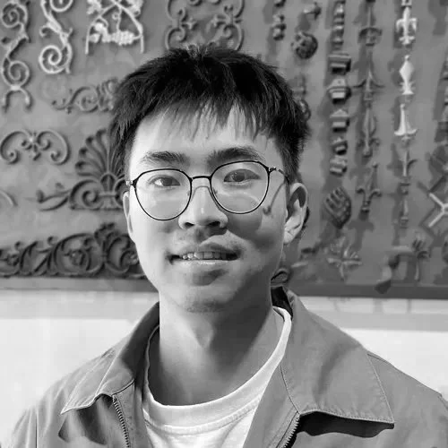 A young man with short black hair, glasses, and a light-colored jacket smiling at the camera, standing in front of a wall decorated with metal sculptures.