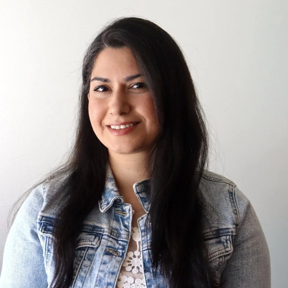 A woman with long wavy hair smiling at the camera, wearing hoop earrings and a beige vest over a navy blue top, standing in front of a colorful patterned tapestry.