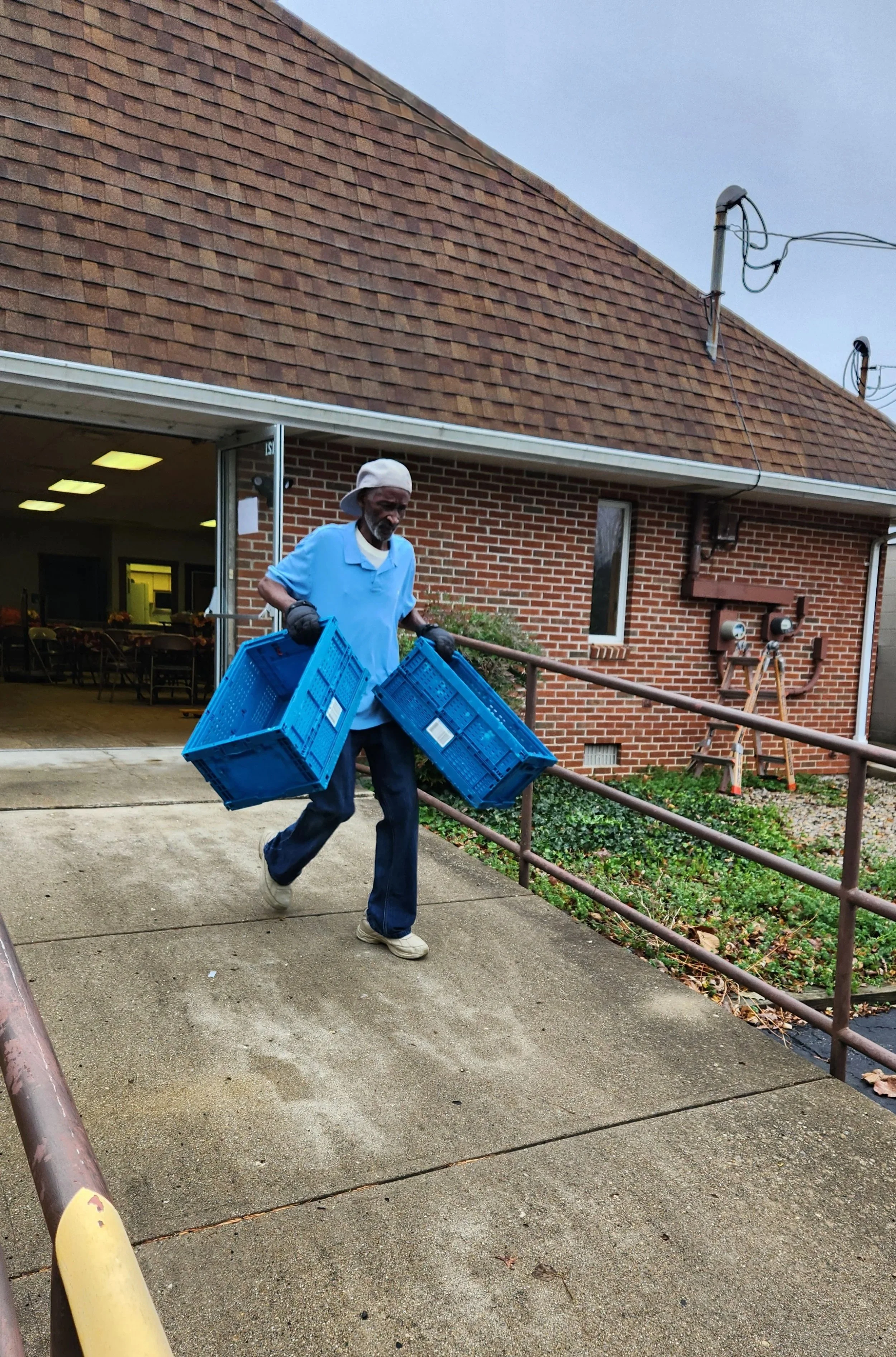 Man carrying blue plastic crates outside a brick building, with an open door leading to a room with tables and chairs. Overcast sky.