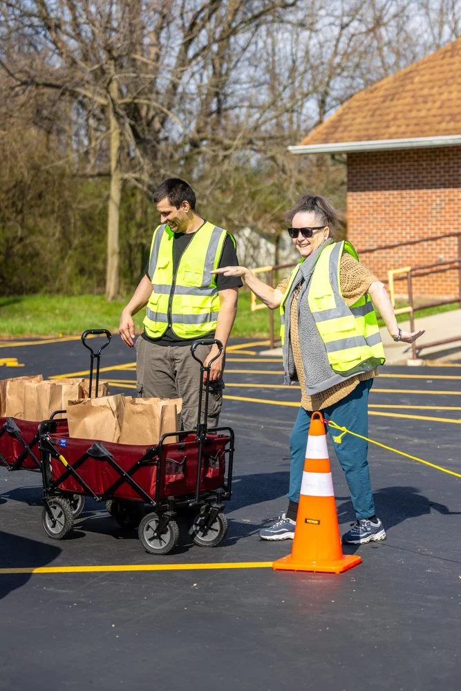 Two smiling people wearing reflective safety vests, standing on a paved lot with yellow lines, next to a orange traffic cone. One person pushes a wagon filled with paper grocery bags, and the other points and smiles. There are leafless trees and a brick building in the background.