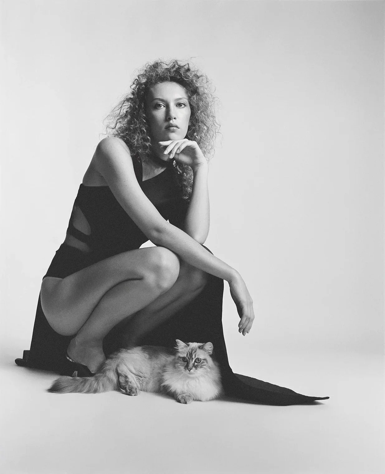 Black and white photo of a woman with curly hair squatting next to a long-haired cat. The woman is resting her chin on her hand, wearing a sleeveless black dress with cutouts, and looking at the camera. The cat is lying on the floor, looking slightly