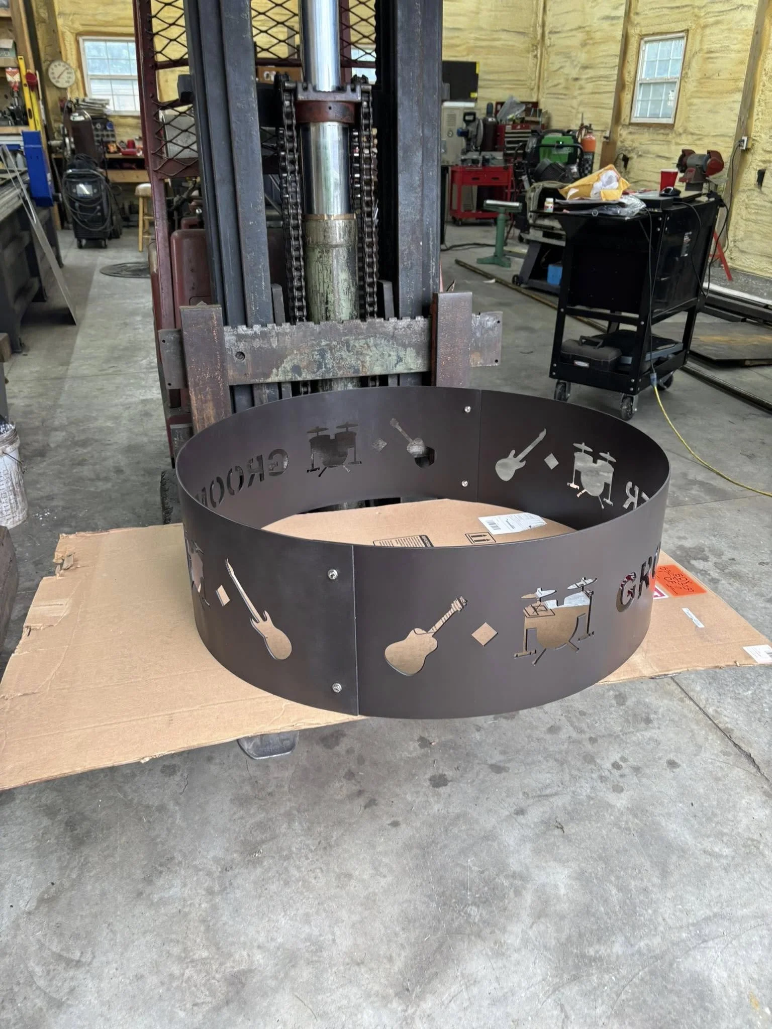 A metal fire pit with cutouts of musical instruments and the word 'Guitar' partially visible, placed on a cardboard sheet in a workshop.