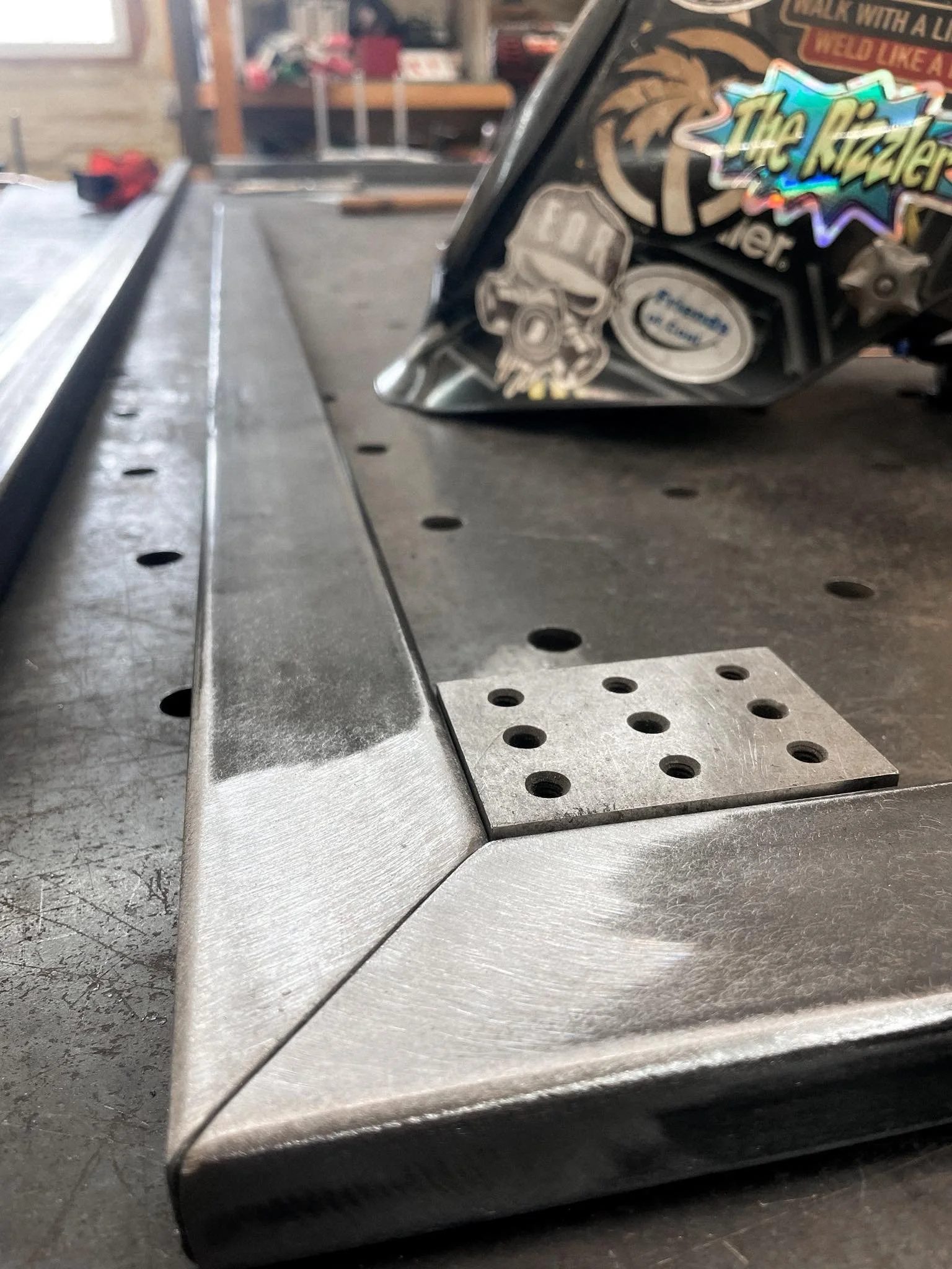 Close-up of a metal workbench with a track, a metal angle with multiple holes, and a pinball machine with colorful stickers in the background.