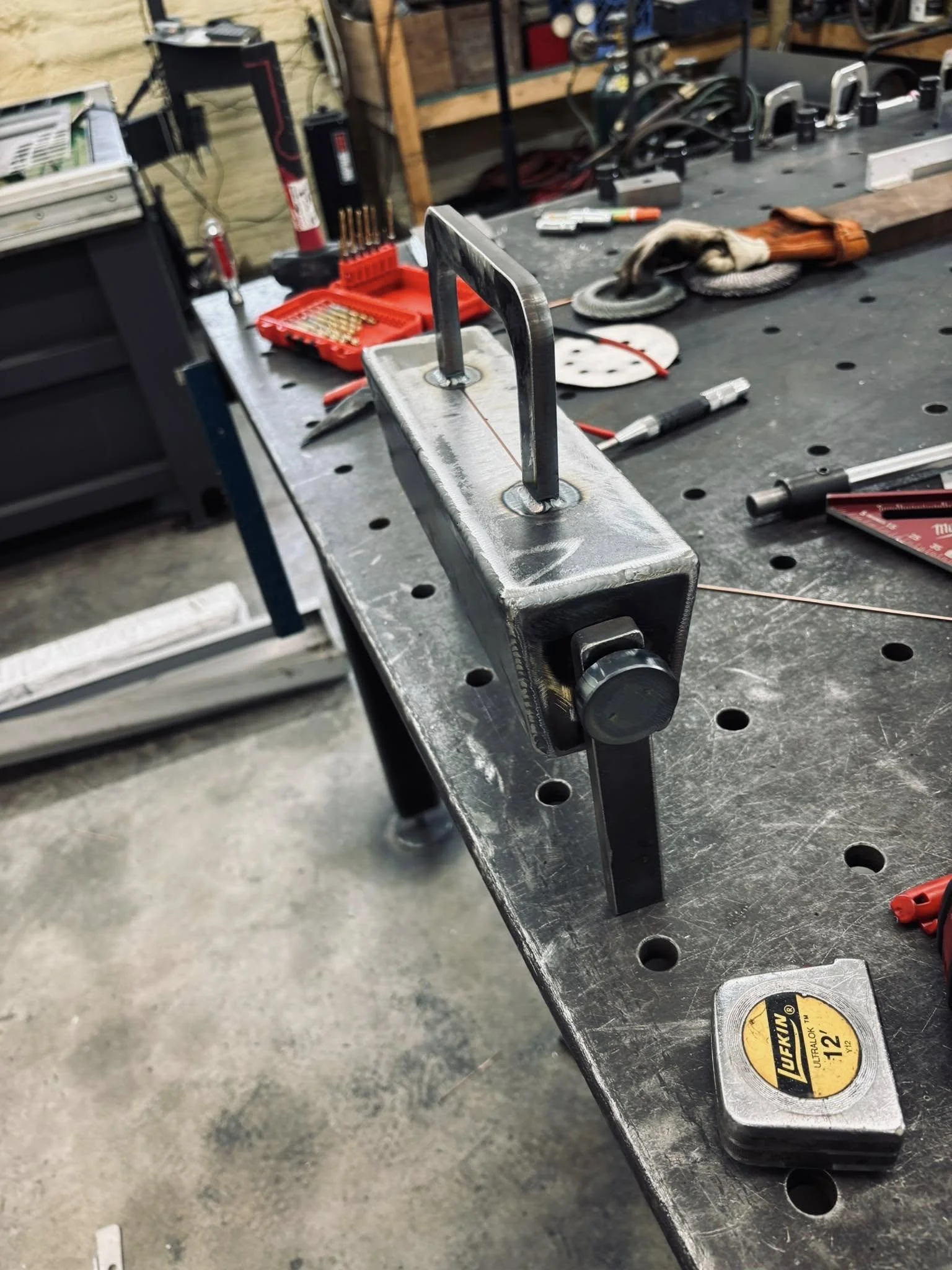 A metal workbench with various tools and equipment, including a red toolbox, a measuring tape, and electrical components, in a workshop setting.