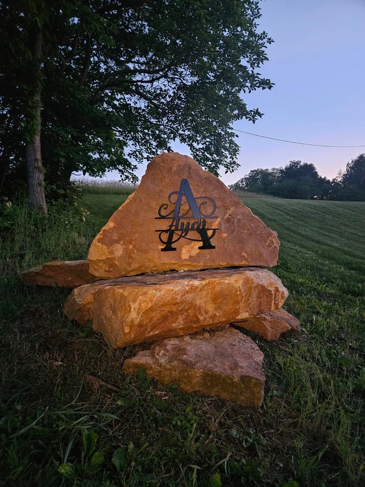 A large carved stone sign with the Audi logo and name, placed outdoors on grass with trees and a sloped hillside in the background during twilight.