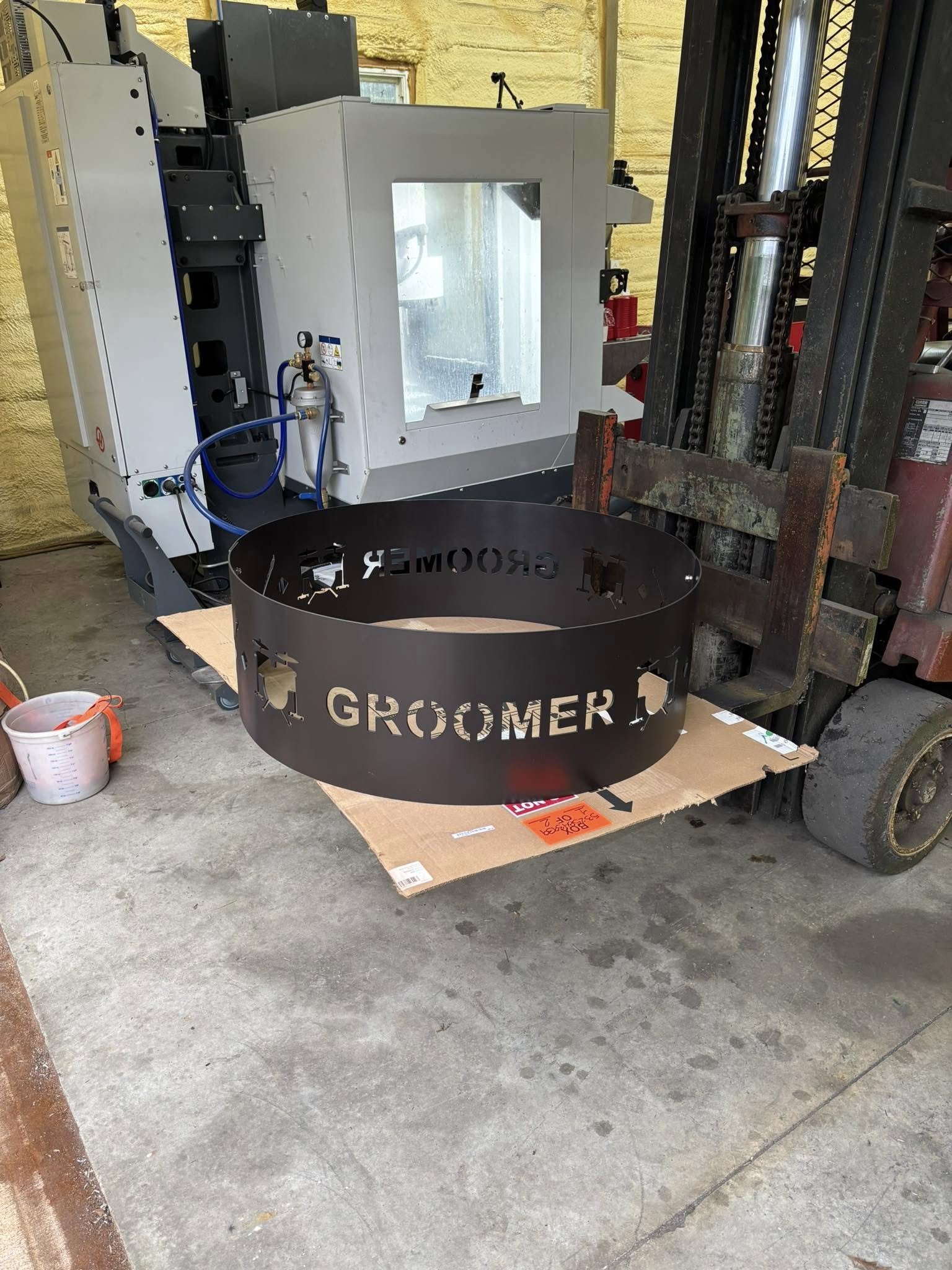 A metal band with cut-out text and icons resting on a piece of cardboard, a forklift, and a machine in the background in a workshop or garage.