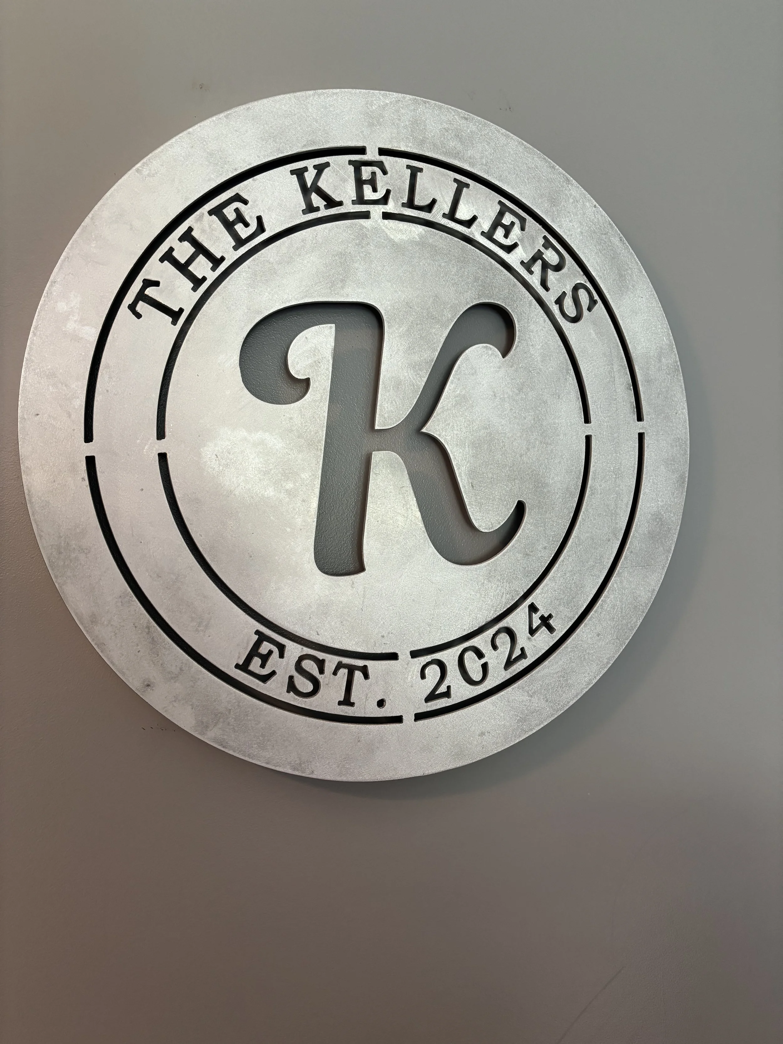Round metallic sign with a large cut-out 'K' in the center. Encircling the 'K' is the text 'THE KELLERS' at the top and 'EST. 2024' at the bottom.