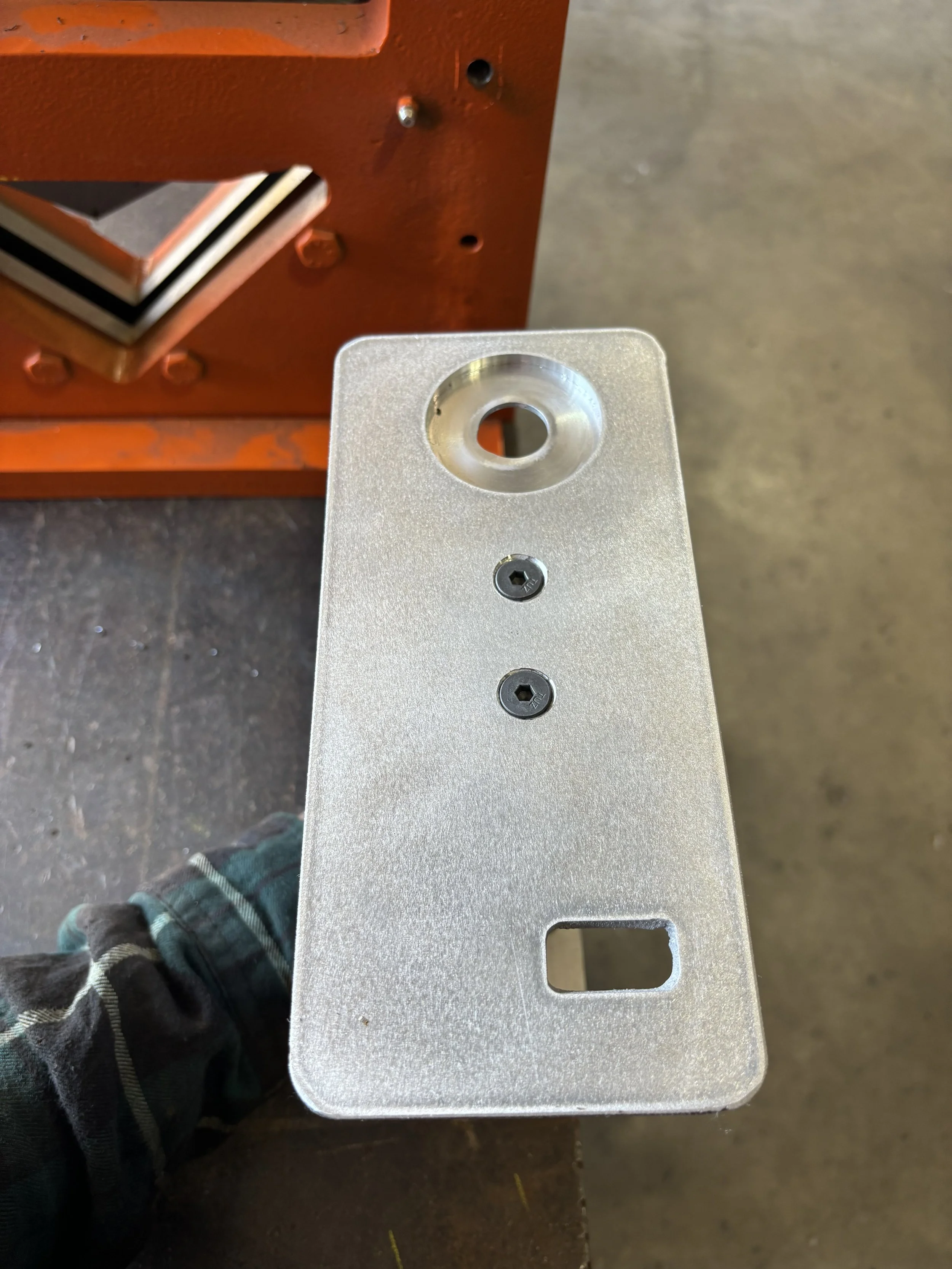 A metal workpiece with a circular hole, two screws, and a rectangular cutout, held in a person's hand wearing a flannel shirt.