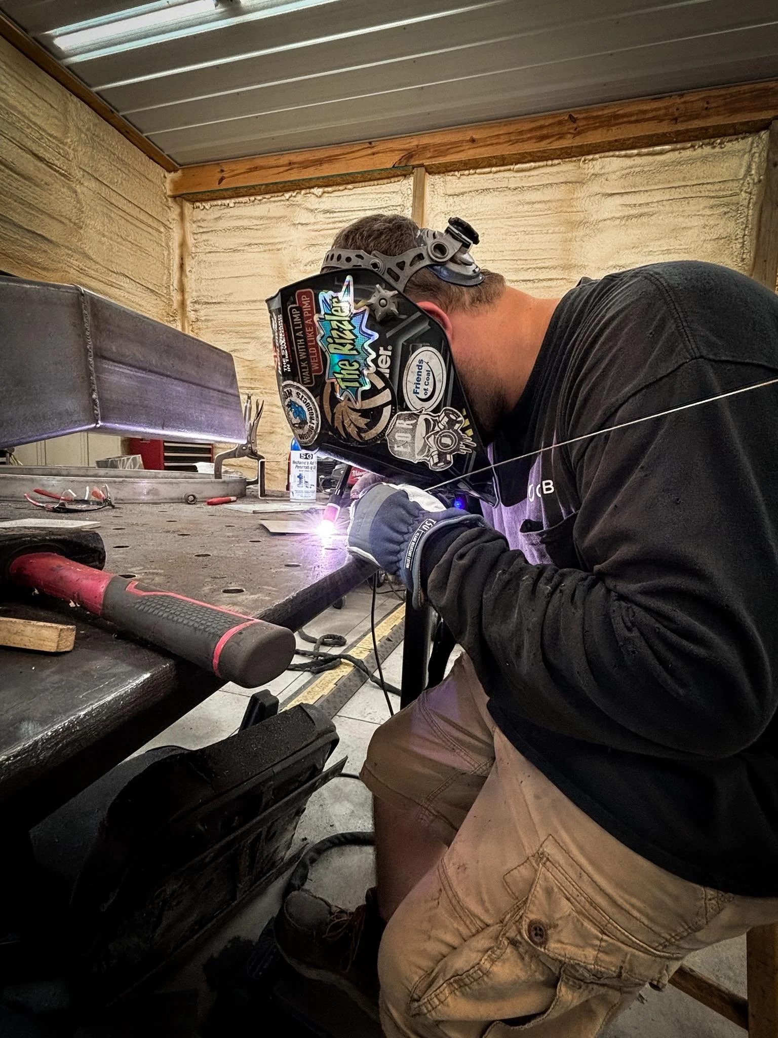 A person welding metal in a workshop, wearing a welding helmet and gloves, with welding equipment on the workbench.