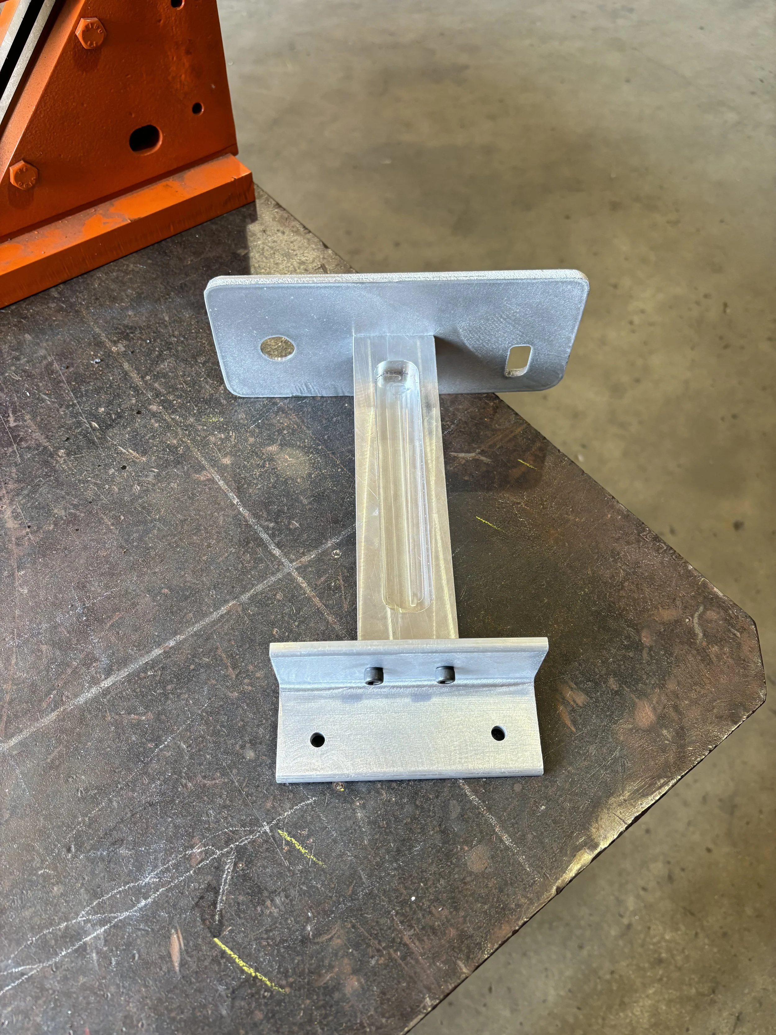Metal bracket with a vertical slot, drilled holes, and a right-angled bend, placed atop a worn, brown work surface with visible scratches and a faint white chalk marking. Part of an orange-colored machine or structure is partially visible in the top-