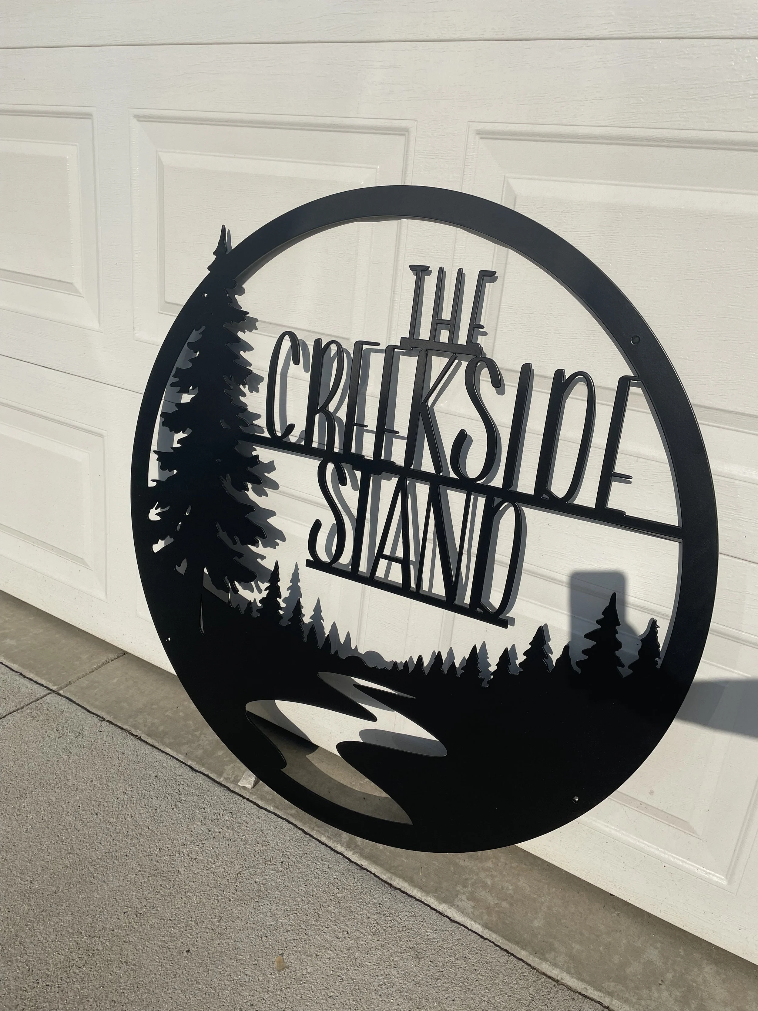 A black metal circular sign with trees and a river cutout, reading 'The Crickside Stand,' leaning against a white garage door.