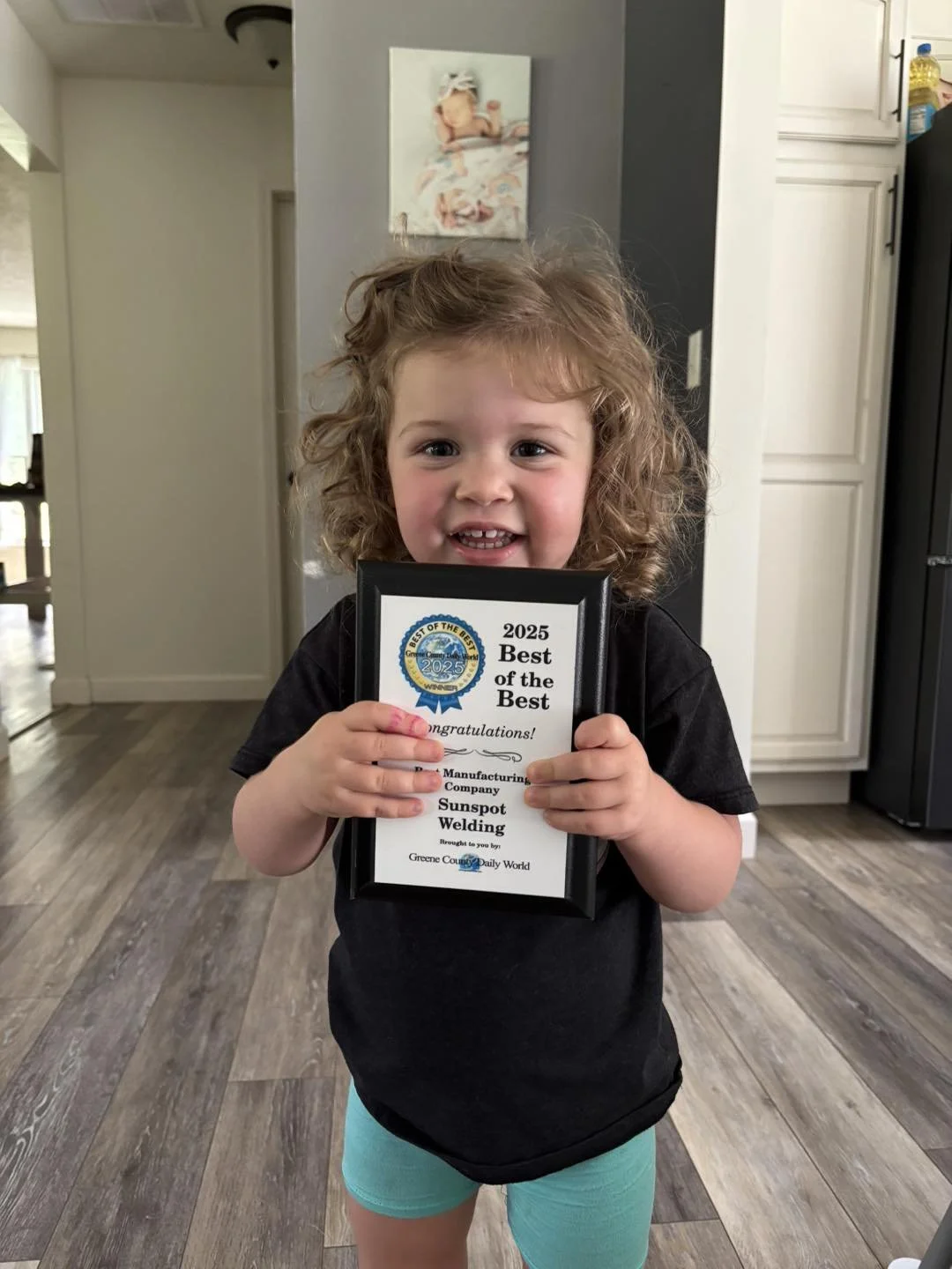 A young girl with curly hair, smiling, holding a framed certificate that says "2025 Best of the Best." She is standing in a kitchen with a gray wall, white cabinets, a black refrigerator, and a wooden floor.