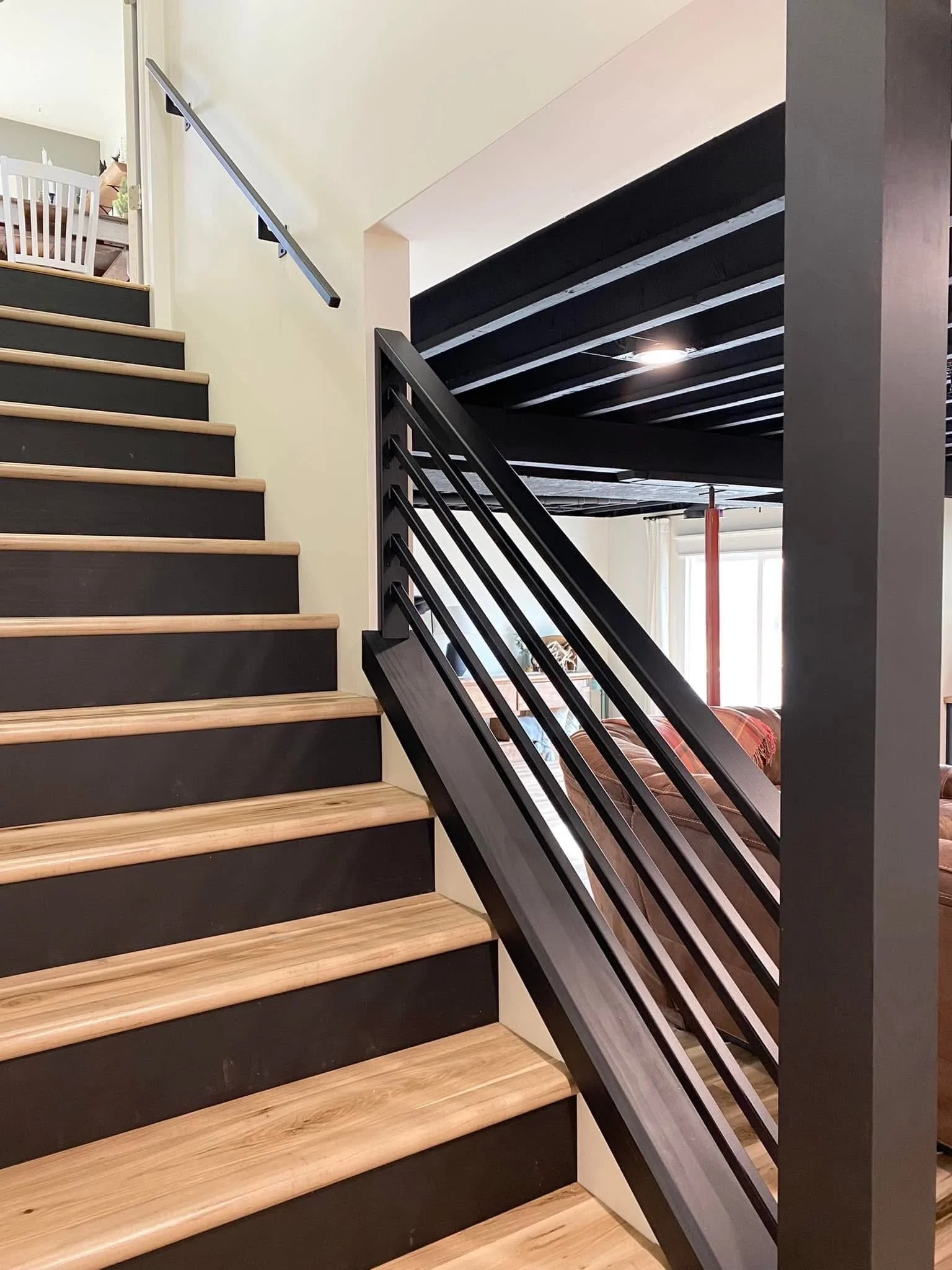 Interior view of a staircase with light wood steps, black risers, and a black metal handrail and balustrade.