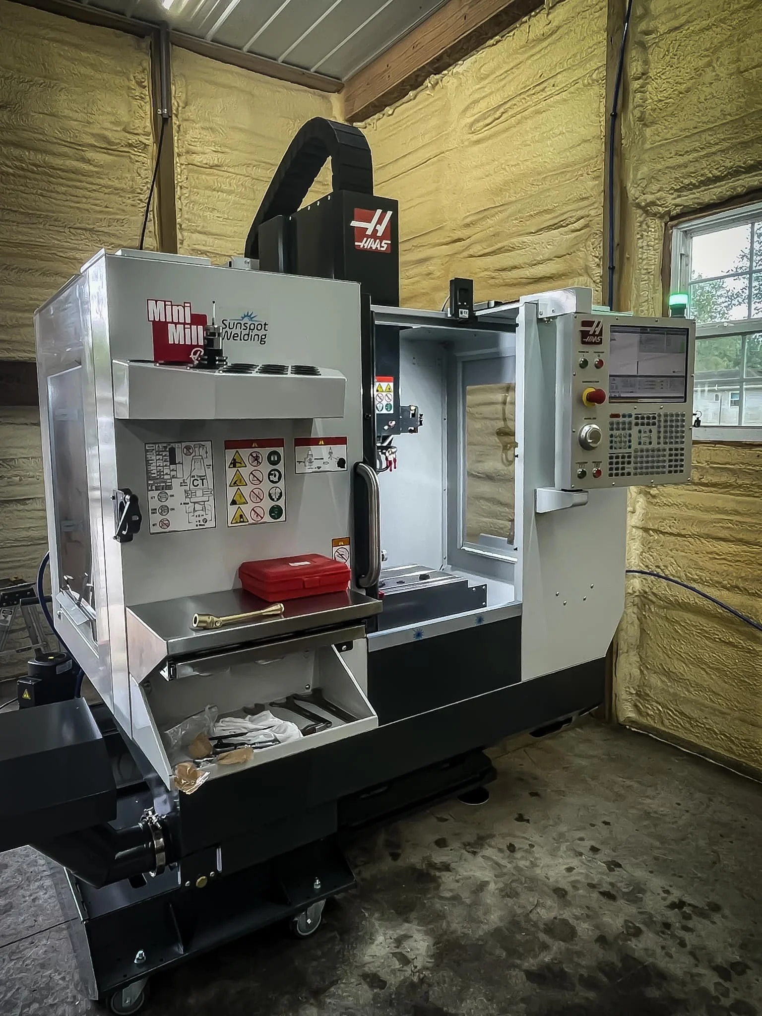 A CNC machine in a workshop with yellow insulation walls and a window, featuring a control panel on the right side.
