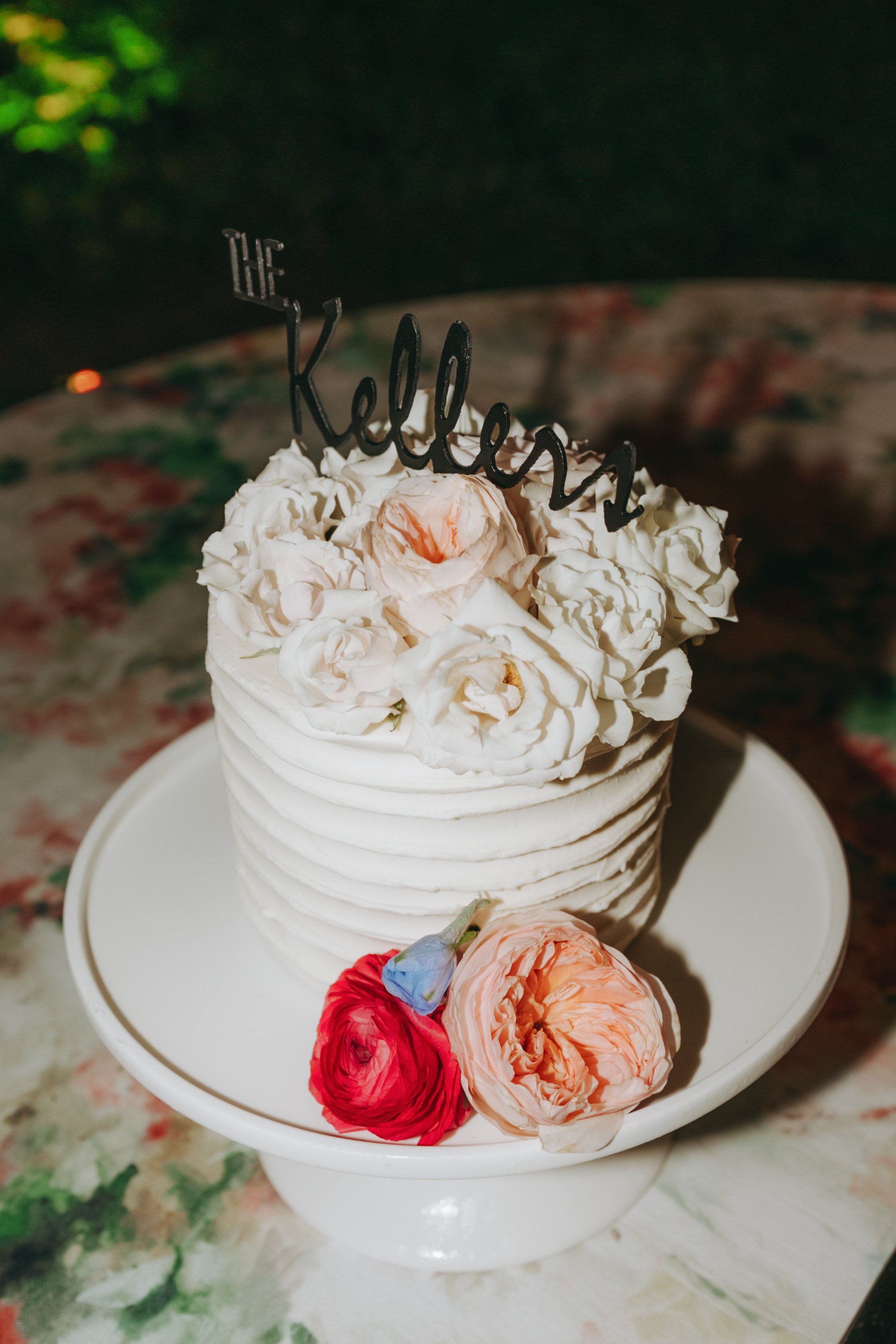 A white cake decorated with roses and a topper that says 'The Kelleys' on a white cake stand.