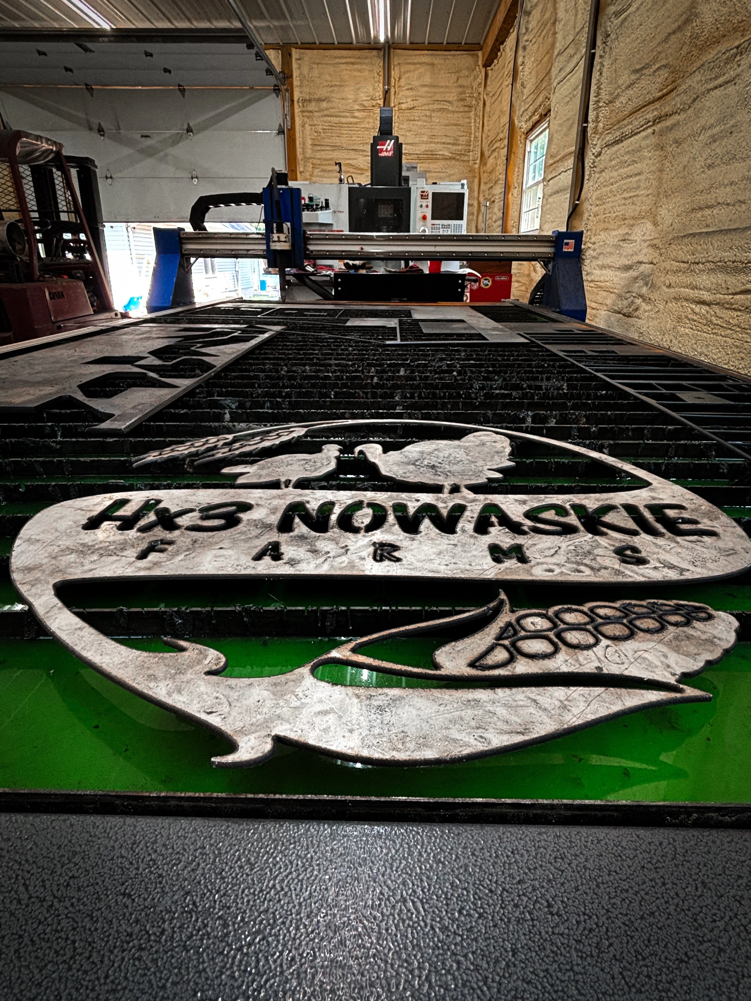 A metal sign with a fish graphic and the text 'HXS NOWASKIE' resting on a green work surface in a workshop. A CNC machine and workshop interior are visible in the background.