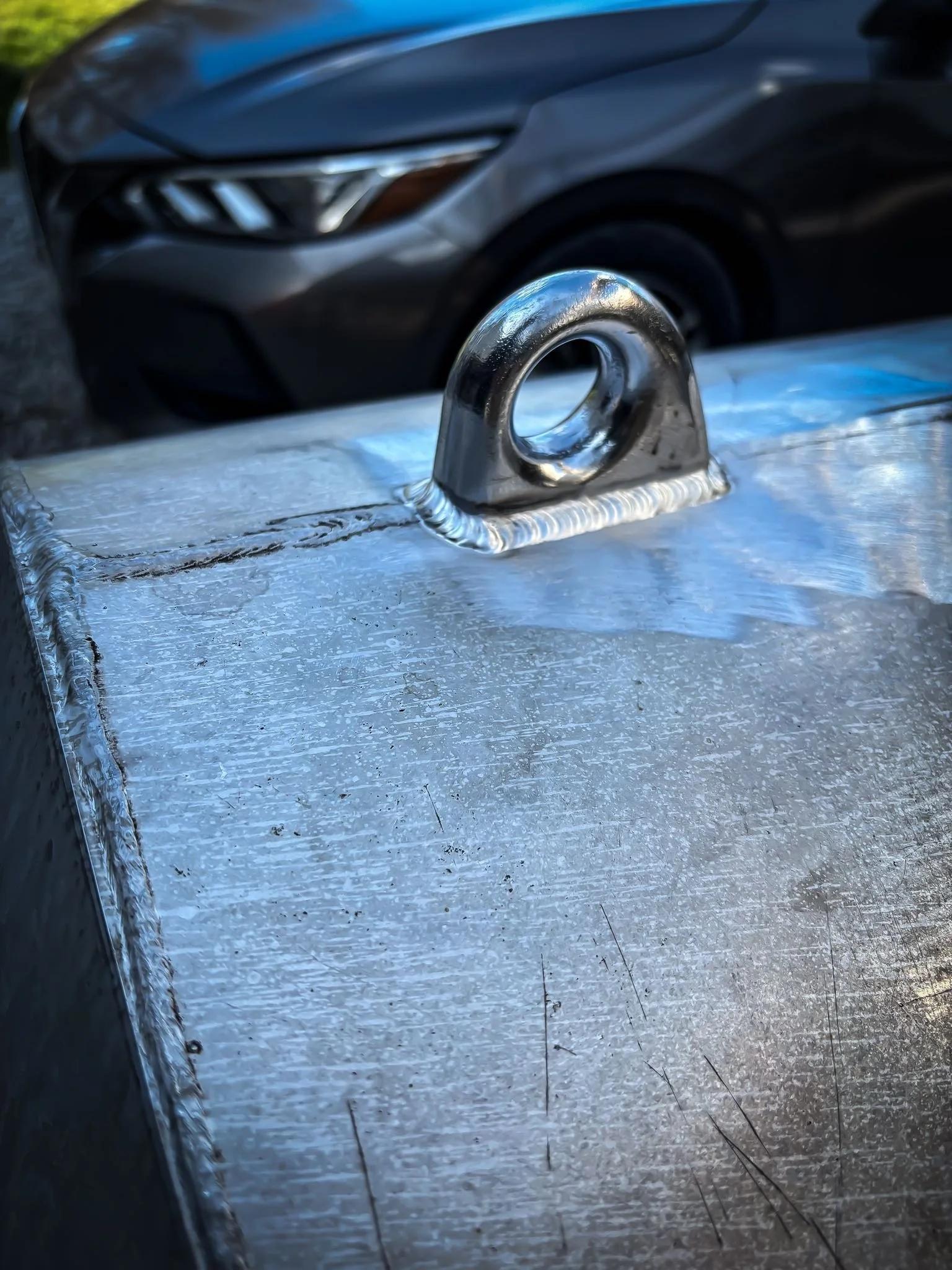 Close-up of a metal eyelet welded onto a surface, with a blurry car in the background.