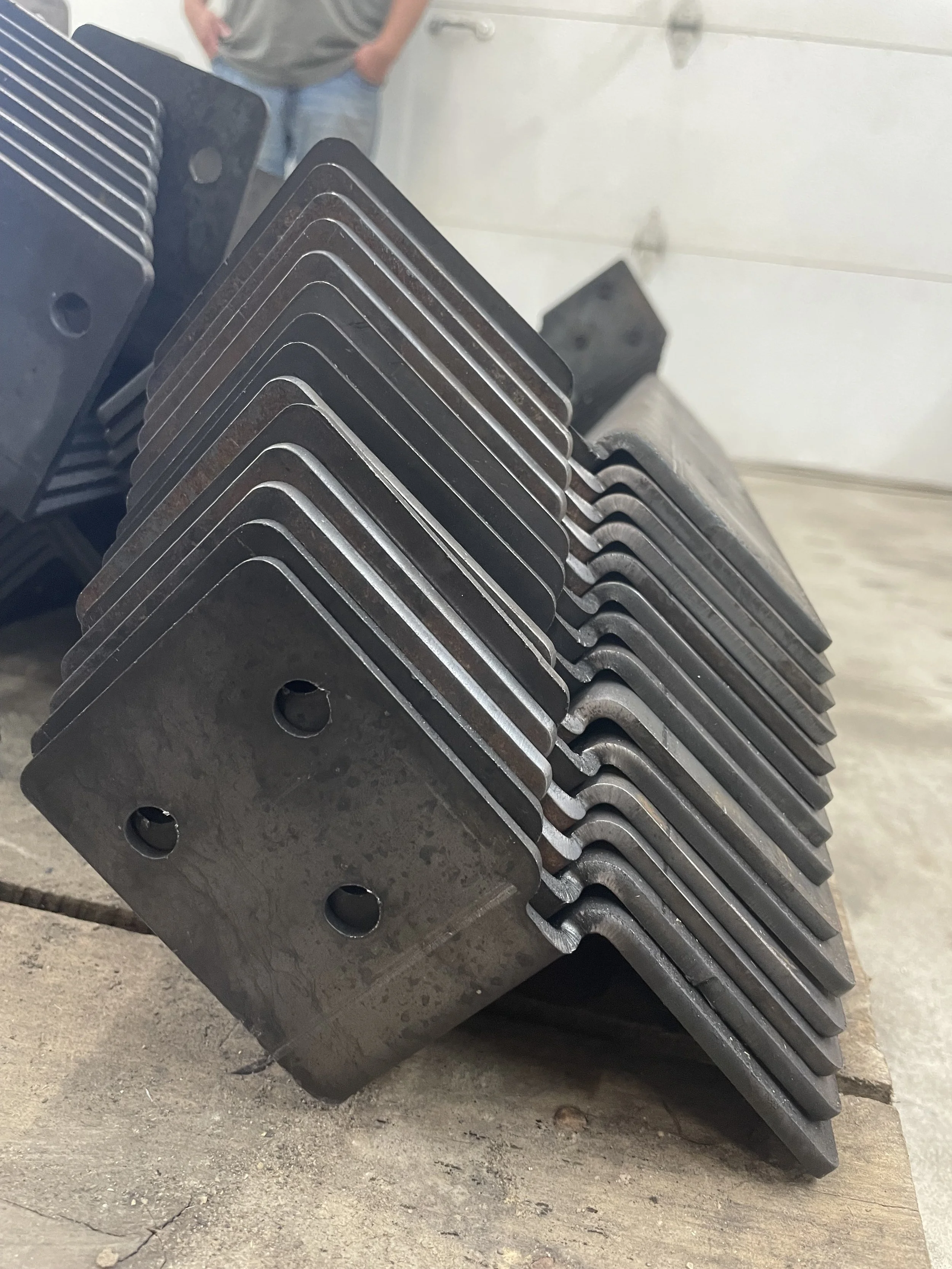 A stack of metal engine or machinery parts with rounded edges and holes, placed on wooden planks in a workshop. A person with hands in pockets is visible in the background.