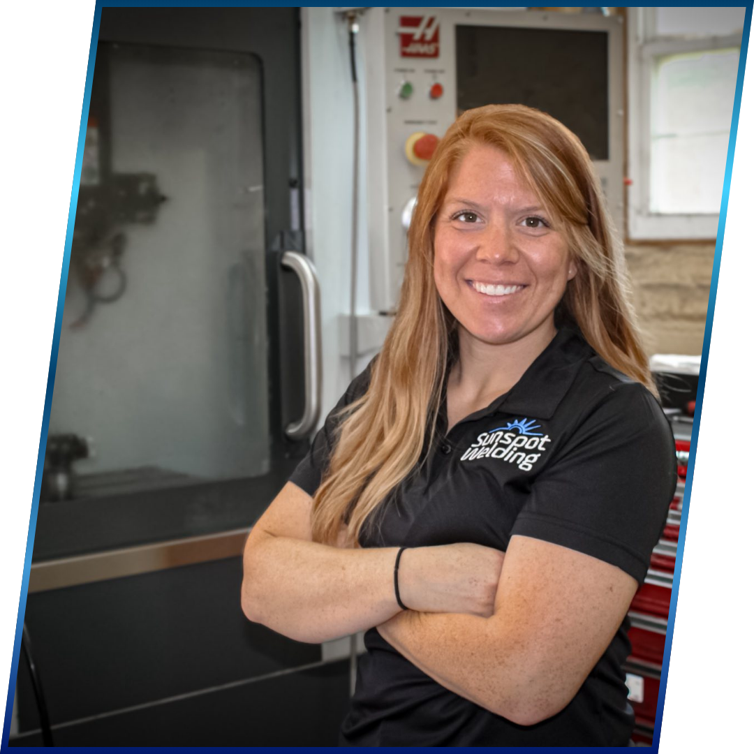 A woman with long red hair smiling and standing with arms crossed in a workspace, wearing a black polo shirt with a logo that reads "Sunspot Welding." In the background, there is a stainless steel oven and a red toolbox.