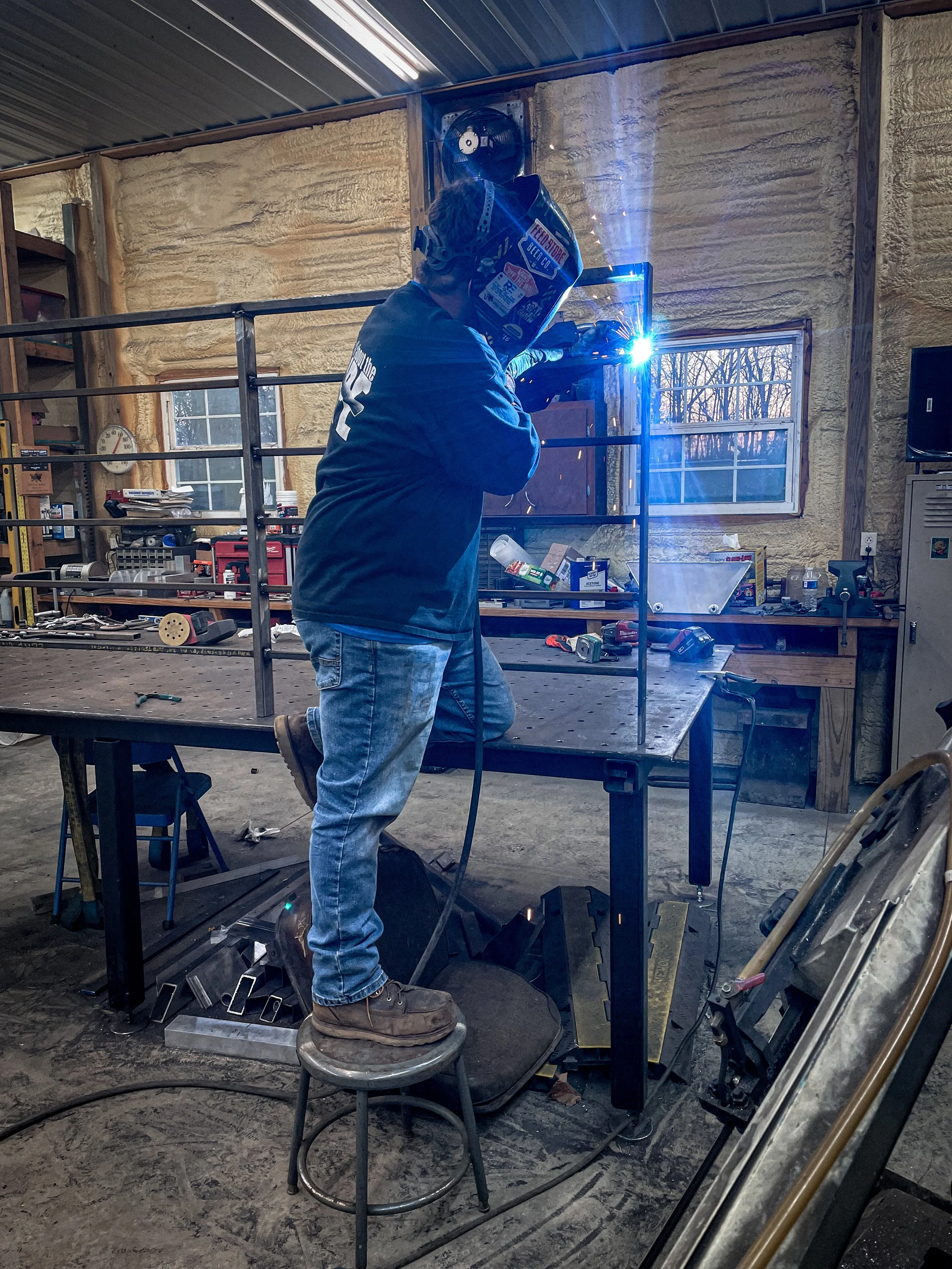 Person welding metal on a workbench in a workshop, wearing a protective welding helmet and gloves.