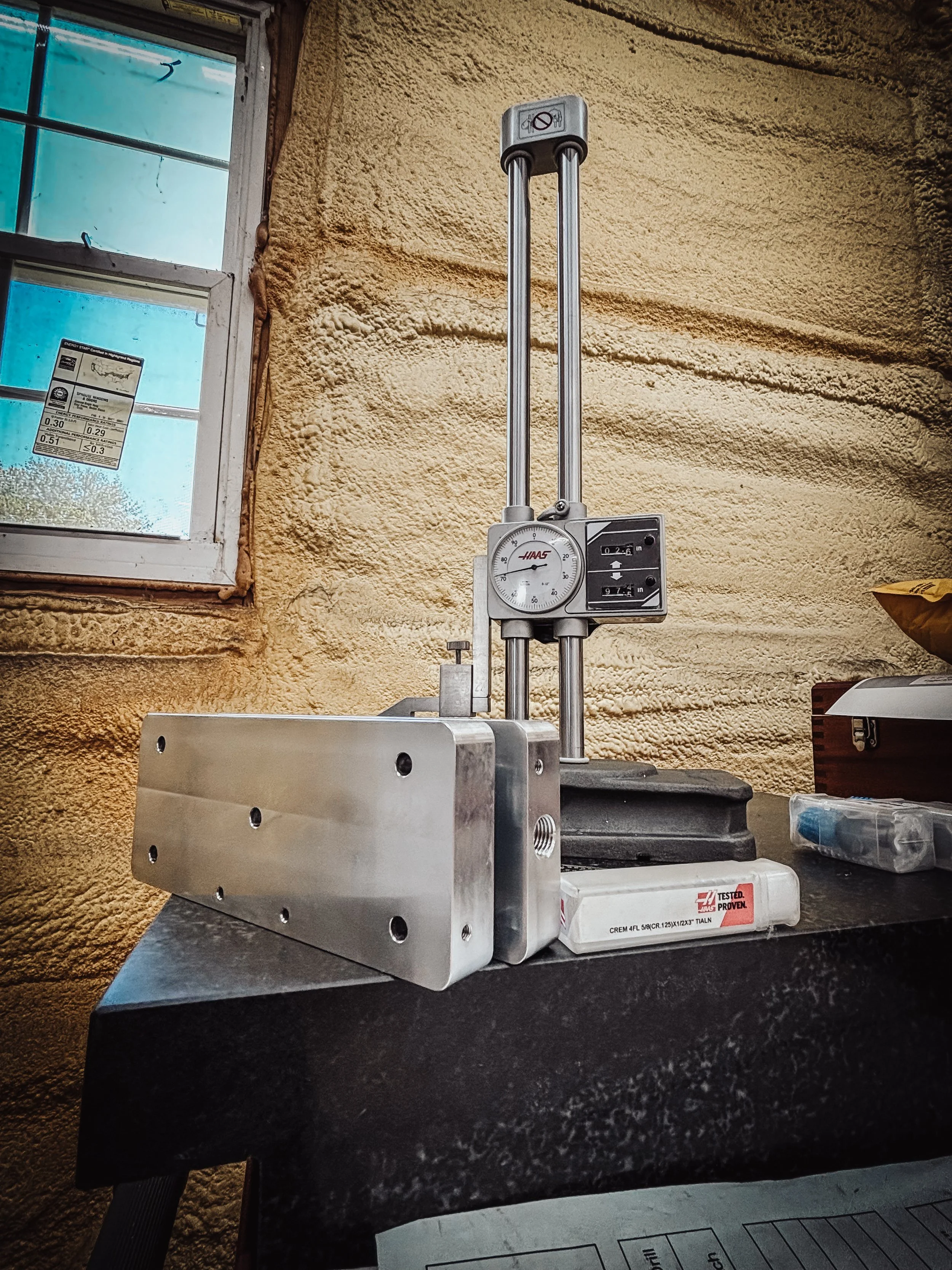 A metallic mechanical device with gauges and rods, placed on a black table in a room with yellow textured walls and a window.