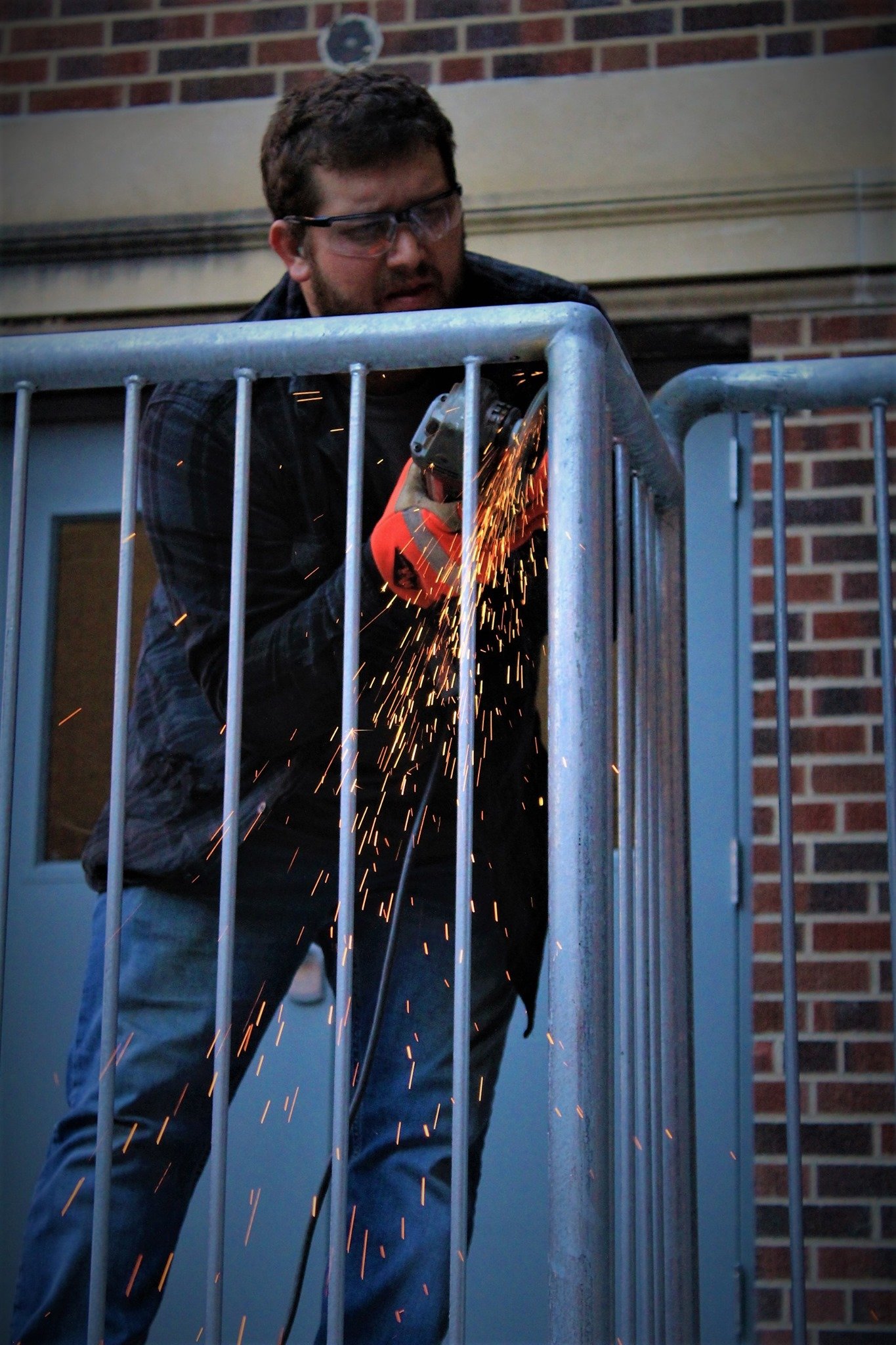 A man wearing safety glasses and red gloves is using a grinder to cut through a metal railing, with sparks flying.