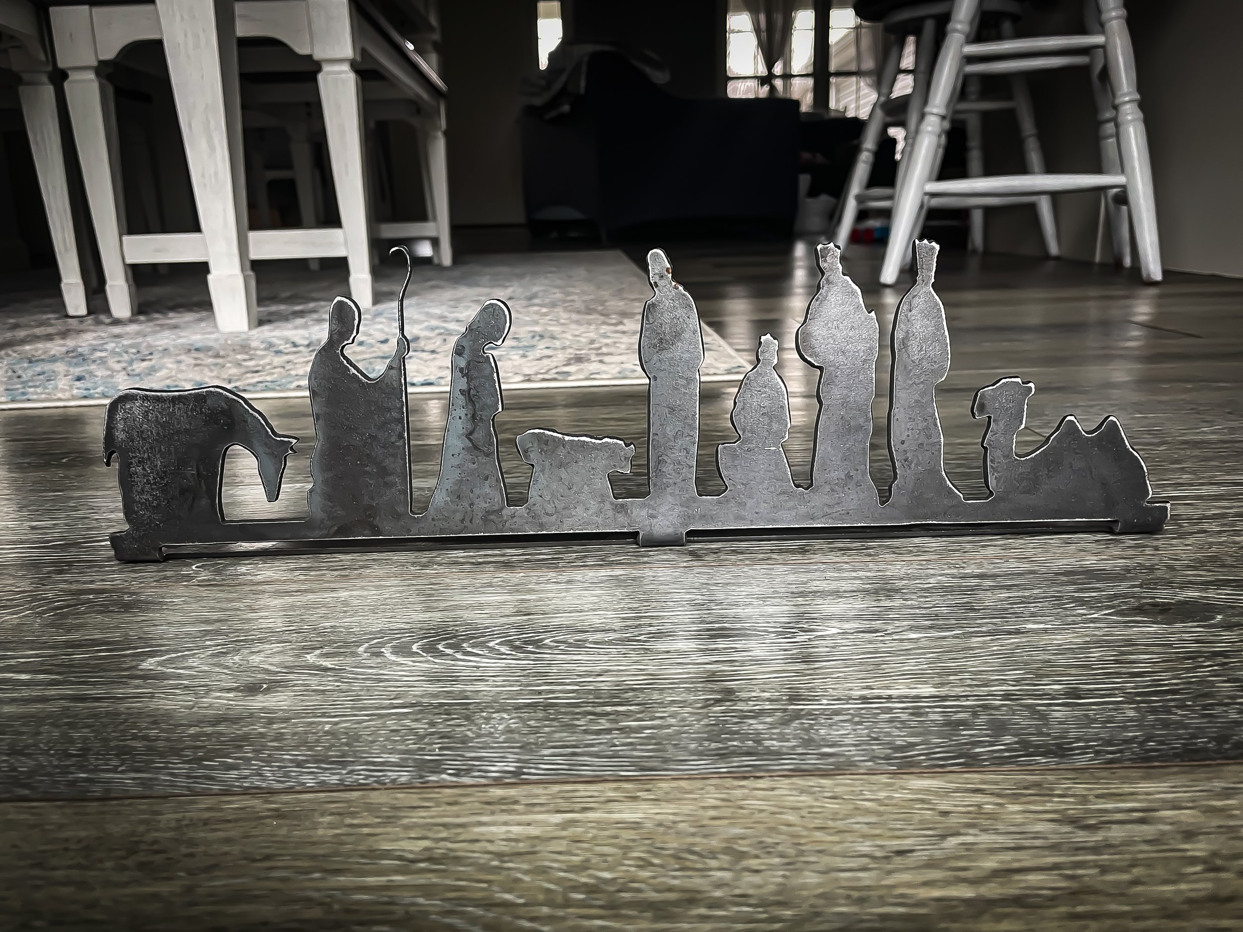 Metal silhouette of a nativity scene with figures including Mary, Joseph, baby Jesus, an angel, shepherds, and wise men on a wooden floor in a dining area.
