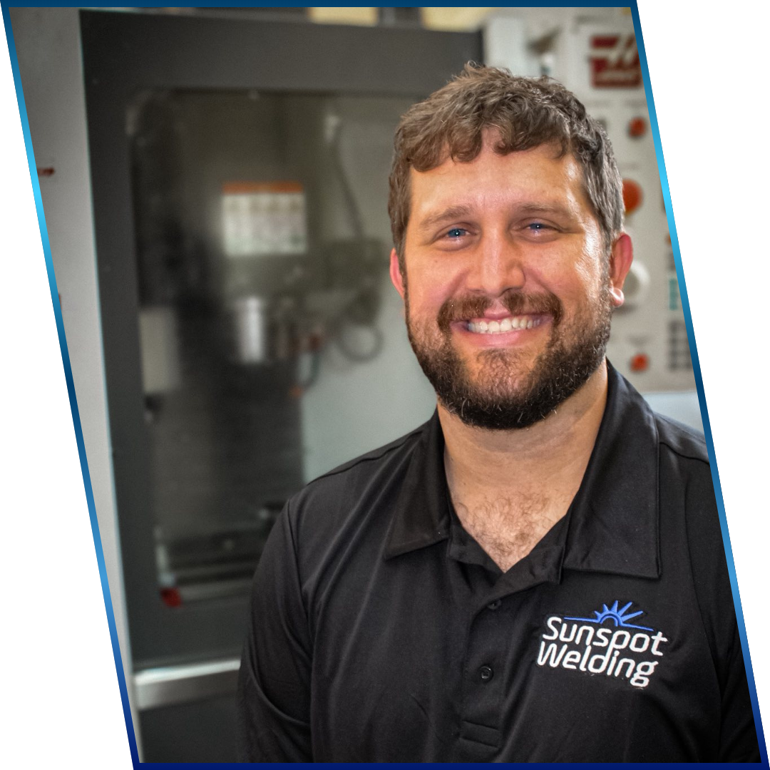 A smiling man with a beard and mustache wearing a black polo shirt with 'Sunspot Welding' logo, standing in front of industrial equipment.