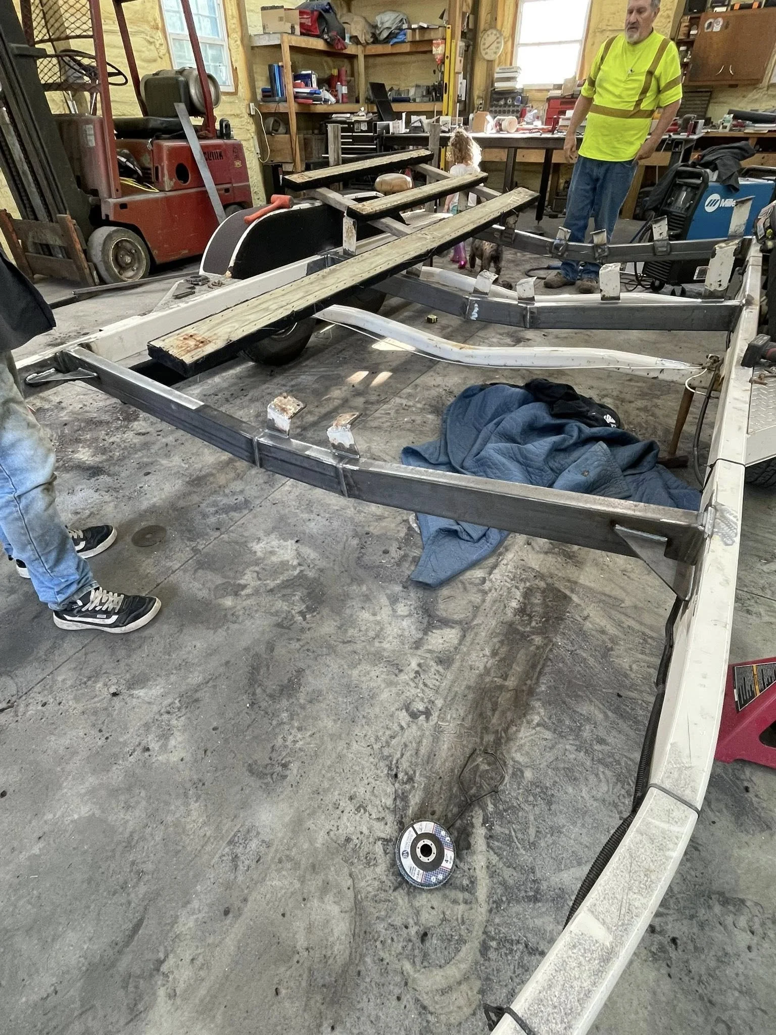People working on a boat frame inside a workshop with tools, shelves, and a dog in the background.