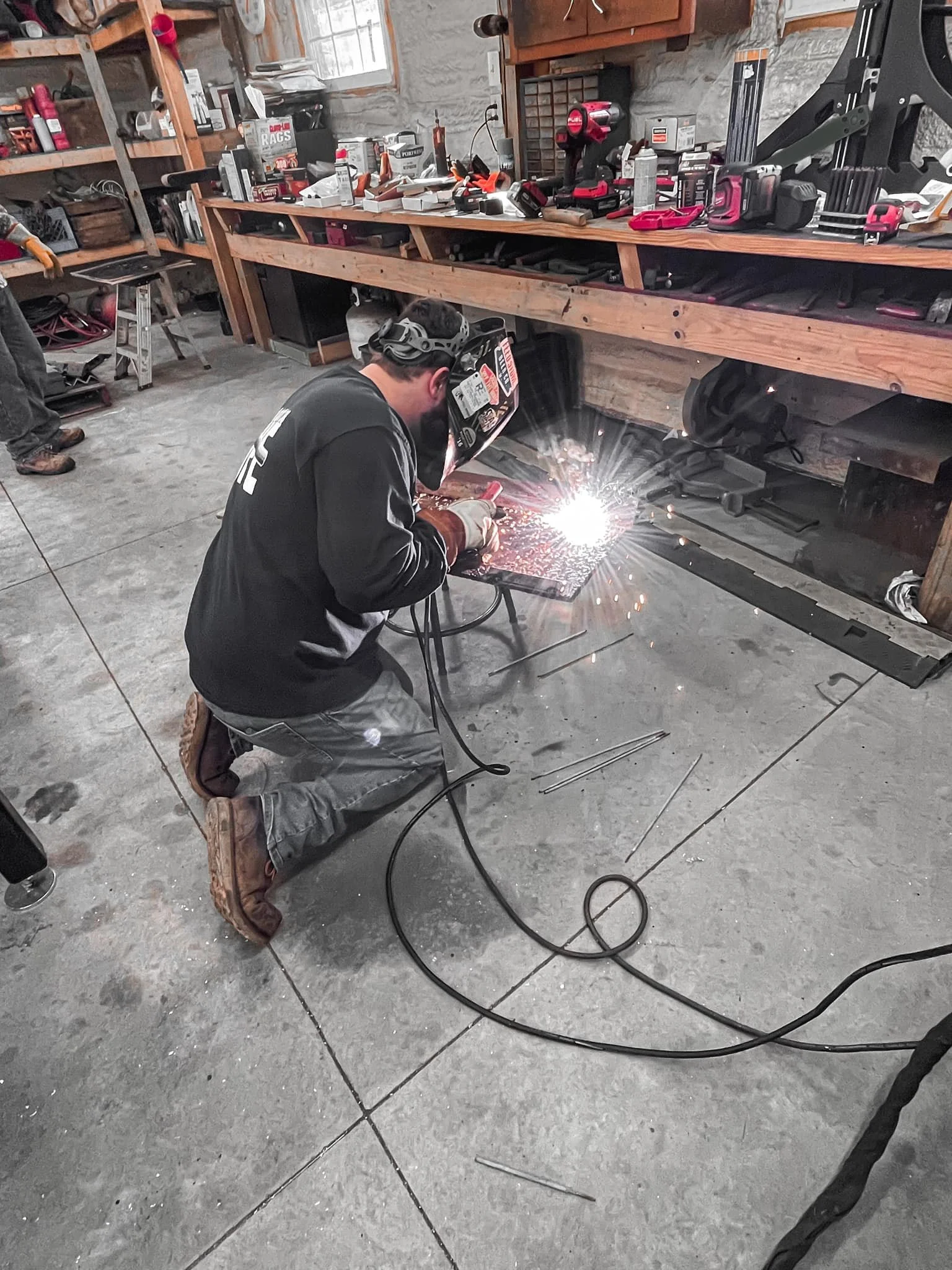 A person welding in a workshop, kneeling on the floor, wearing safety goggles and gloves, sparks flying from the welding process. The workspace has tools and supplies on shelves and a workbench.