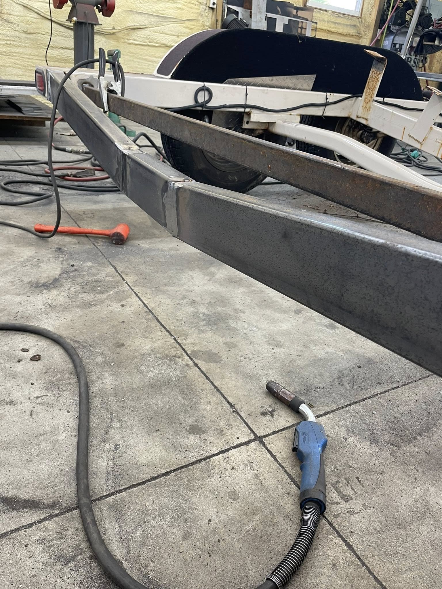 Close-up view of a welding work area showing a metal trailer frame, welding equipment, and floor with scattered tools and cables.