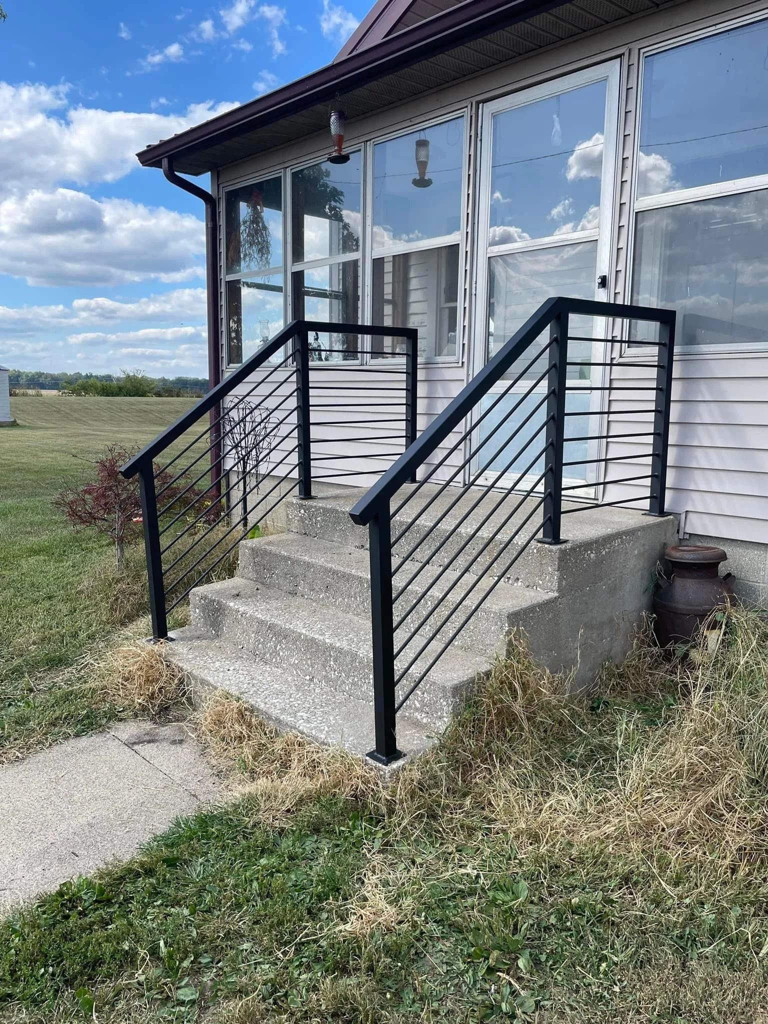 Concrete steps leading to a glass door with black metal railings, attached to a white house with large windows, in a rural area under a partly cloudy sky.