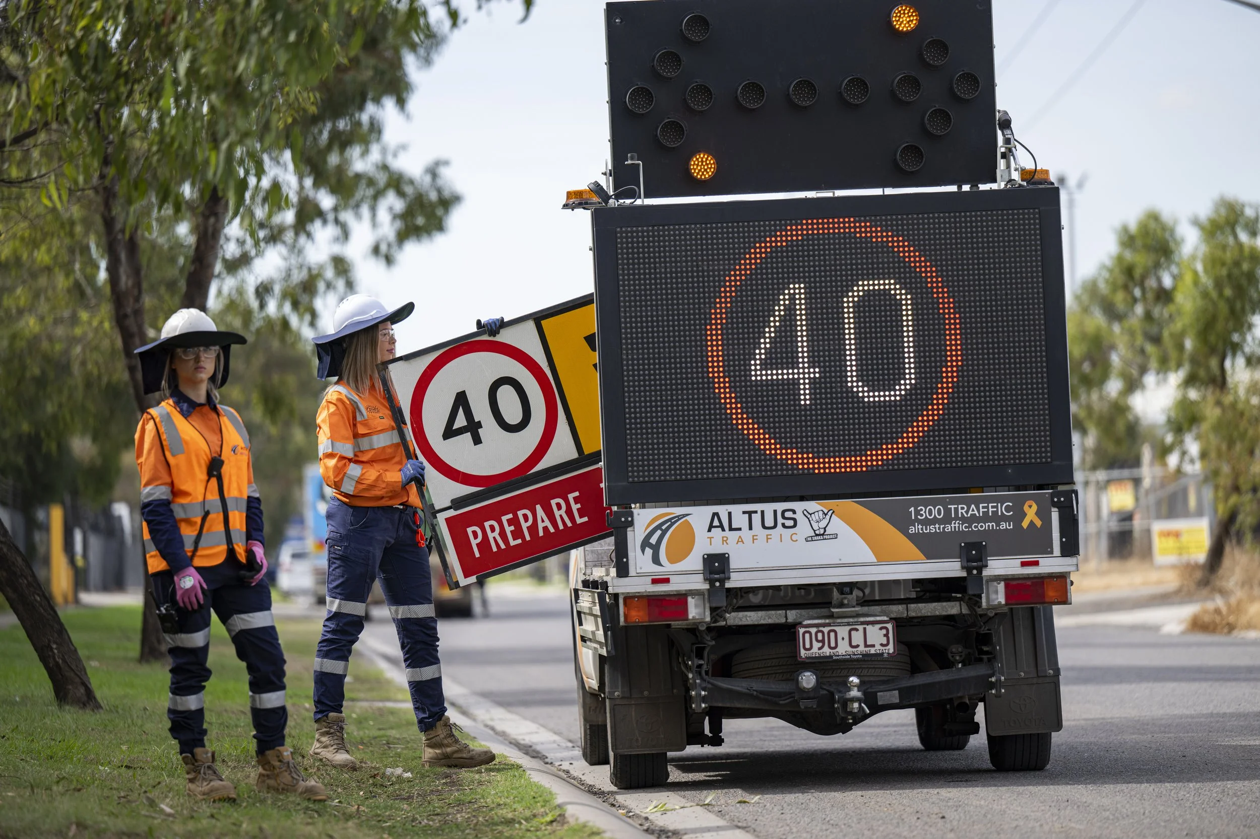 Two workers in orange safety vests and hats standing next to a road sign and digital speed display showing 40 km/h, on a street with trees and a fence in the background.