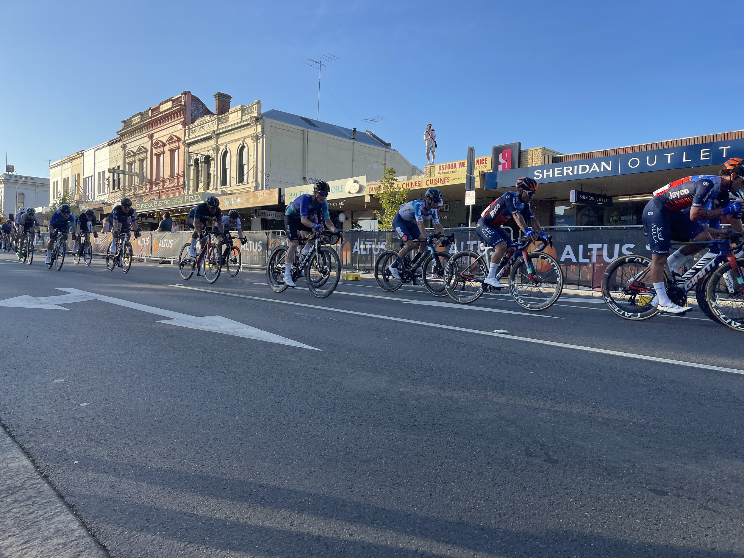 Cyclists participating in a race ride along a city street with storefront signs and a clear blue sky in the background.