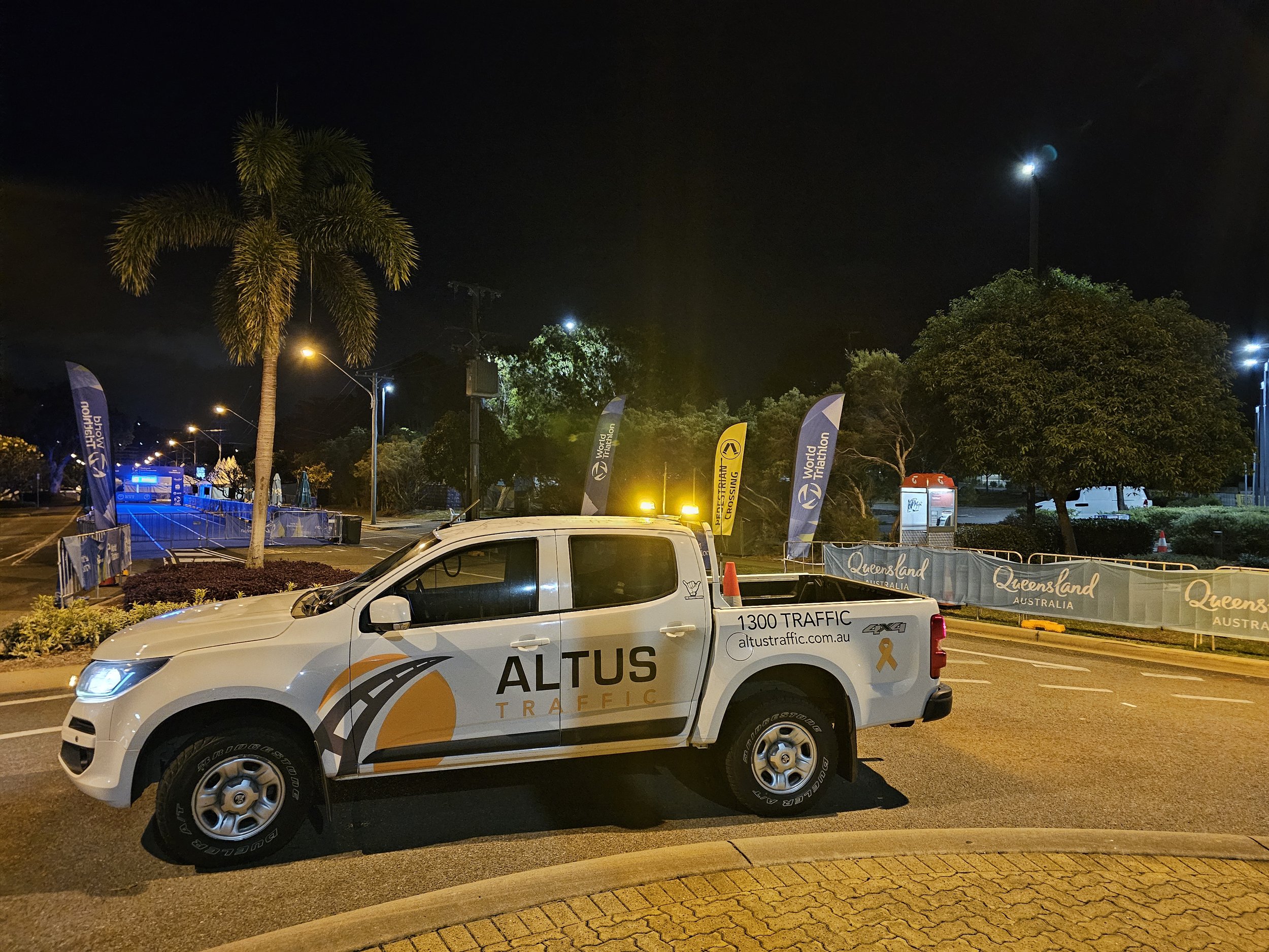 Nighttime scene with a white traffic enforcement vehicle marked 'ALTUS TRAFFIC' parked on the road near Queensland, Australia. The vehicle has a yellow ribbon decal and cones on top. In the background, there are trees, flags with 'World Triathlon' lo