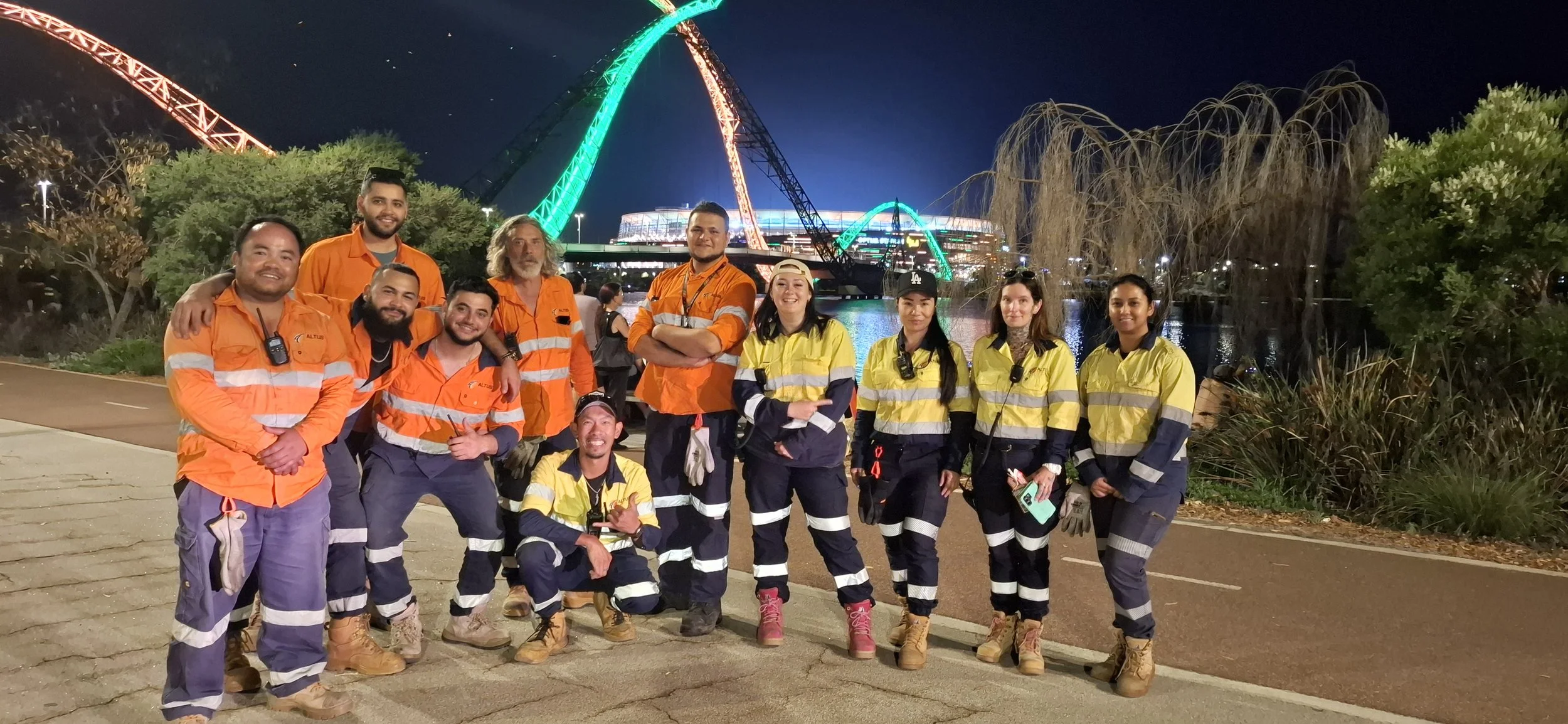Group of eleven emergency response workers in reflective uniforms, smiling, standing on a paved path at night, with a lit ferris wheel and bridge in the background in an urban park setting.