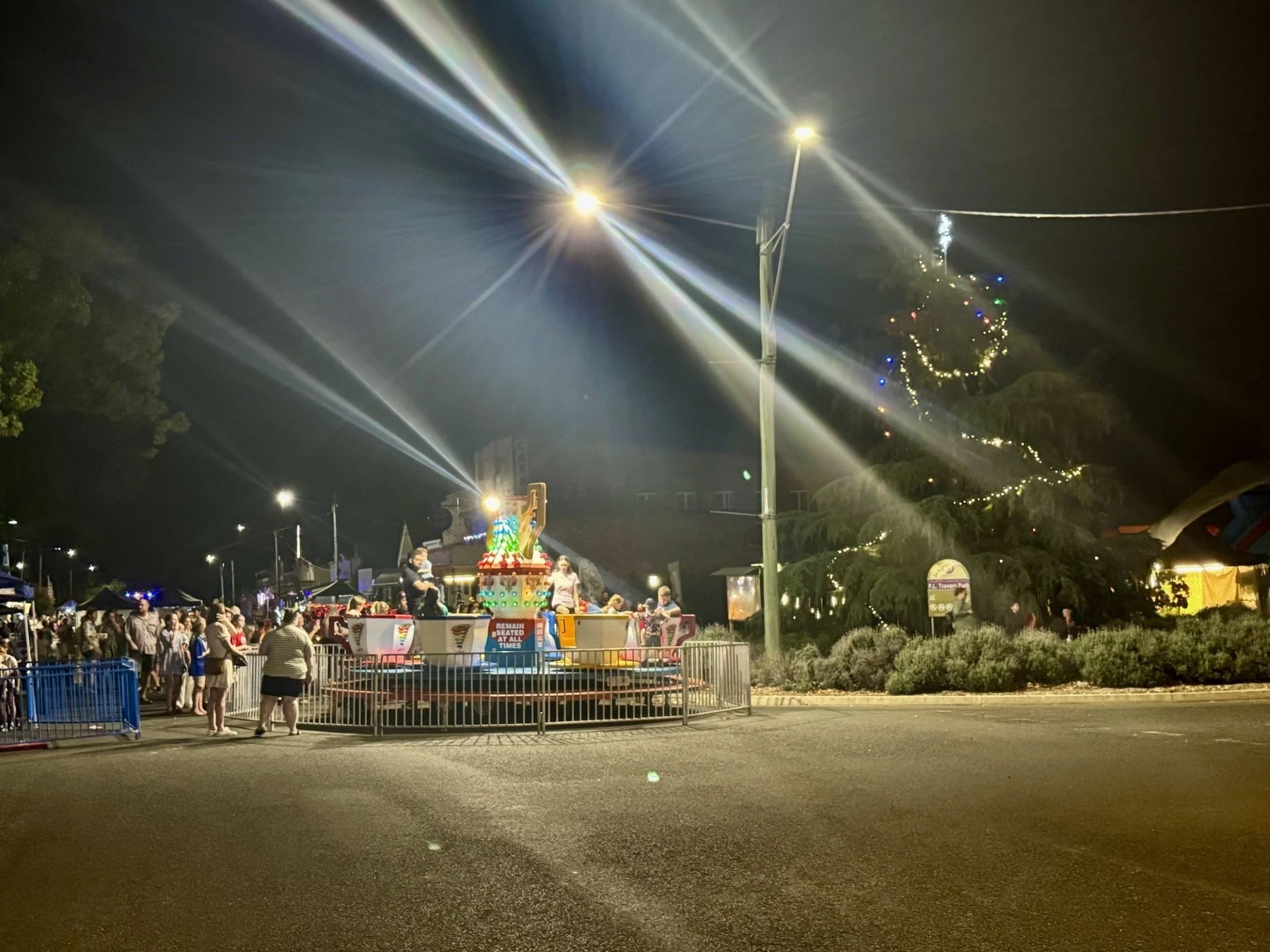Nighttime scene at a carnival with a merry-go-round ride, fireworks in the sky, and a decorated Christmas tree on the right side. People are gathered around the ride, and the area is illuminated by streetlights and colorful lights.