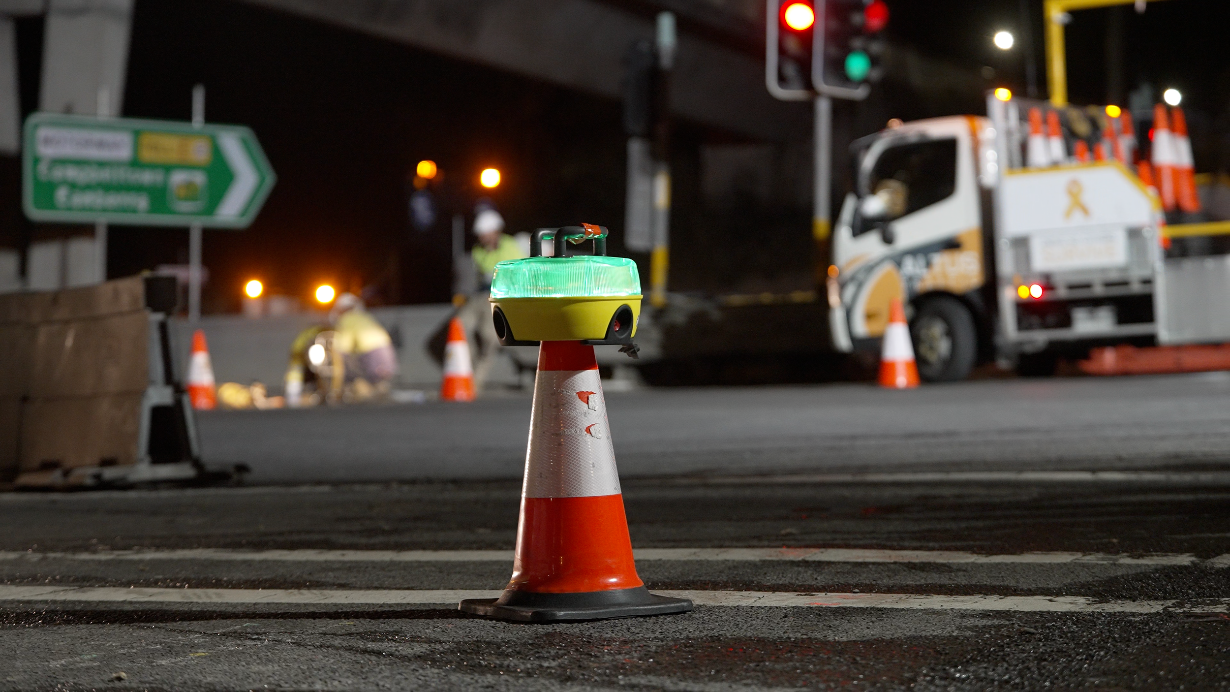 Nighttime road construction scene with orange traffic cone in foreground, illuminated construction equipment, workers, traffic lights, and orange barrels in background.