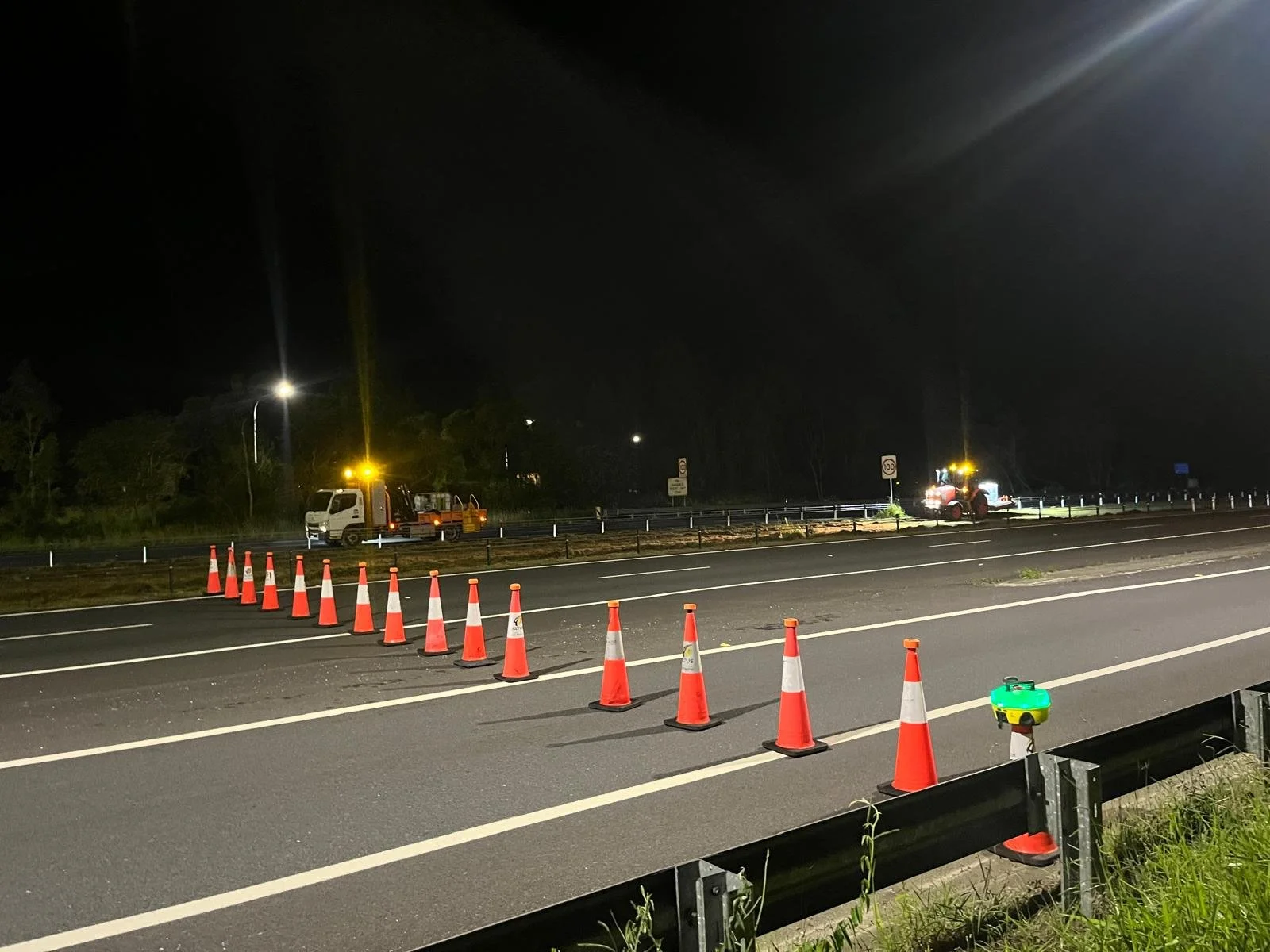 Nighttime road construction scene with orange traffic cones, work trucks, and equipment on a highway.