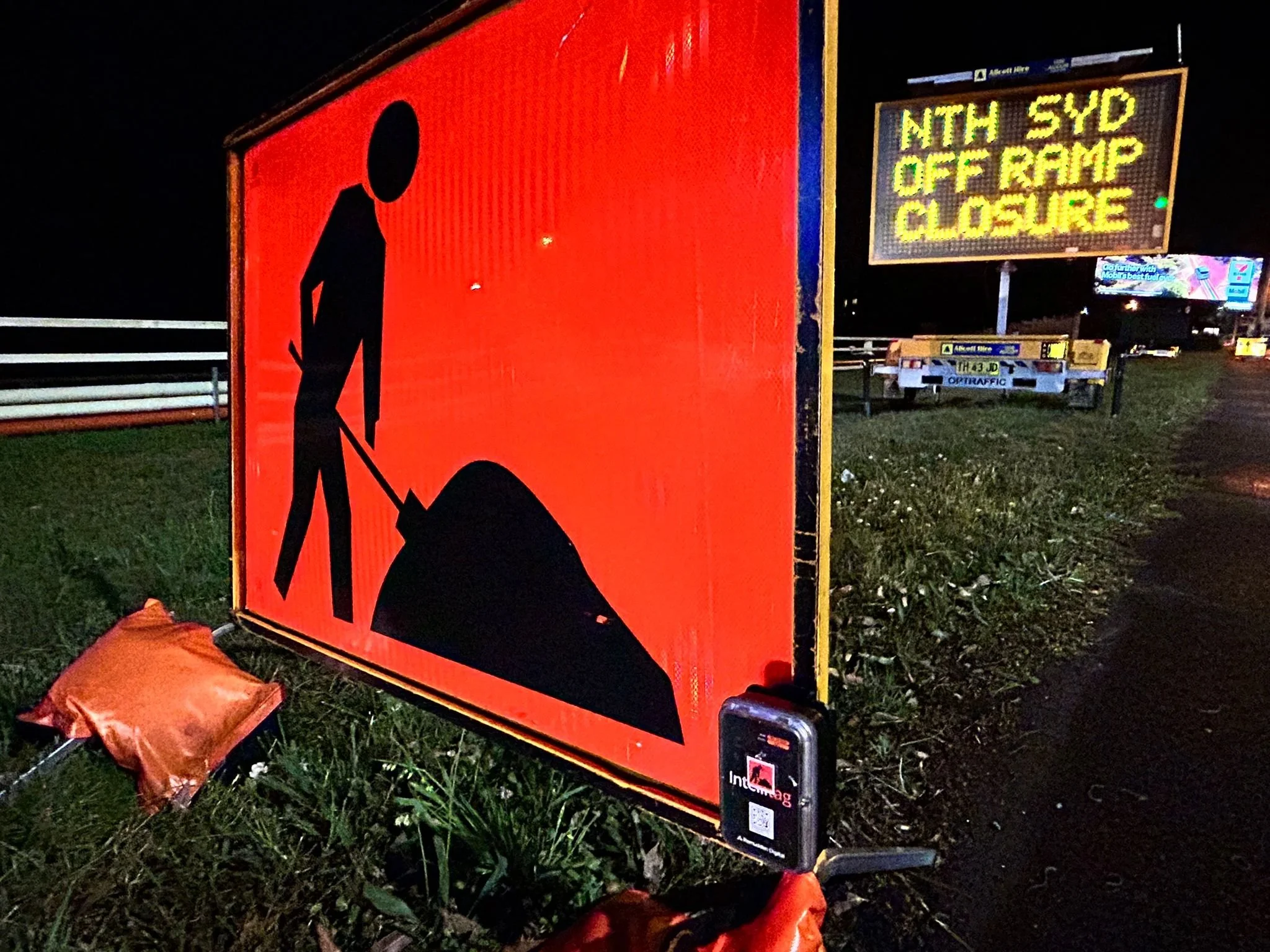 A construction warning sign with a black silhouette of a person shoveling on an orange background. In the background, there is an electronic message board with the text 'NTH SYD OFF RAMP CLOSURE' and a yellow road work vehicle.