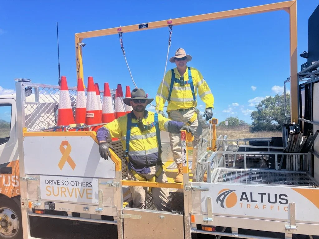Two workers in yellow safety vests and wide-brimmed hats standing on a traffic control vehicle with orange cones and safety signs. One is standing on a platform and the other is sitting, with a clear blue sky and trees in the background.