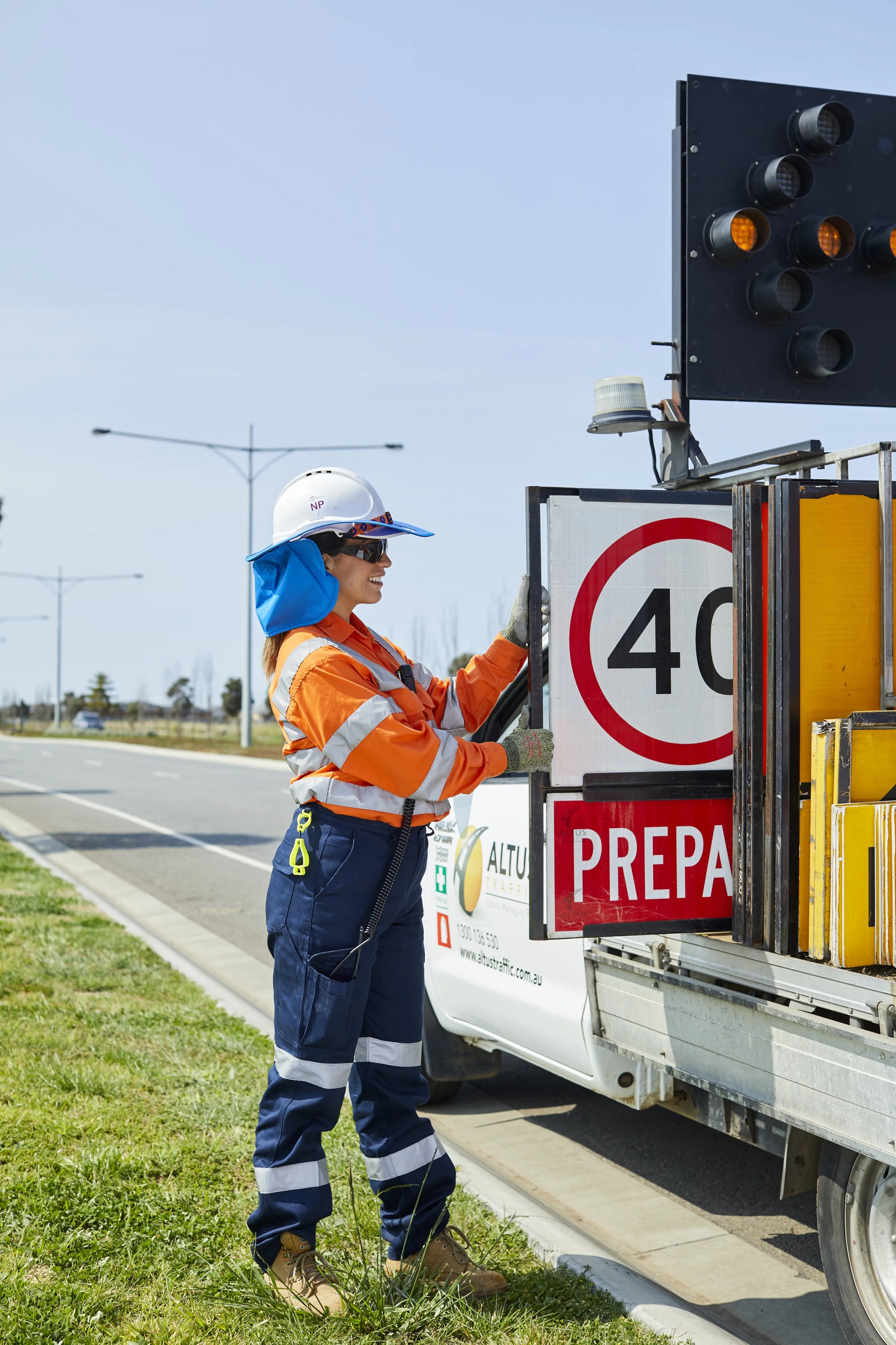 A woman in construction safety gear, including a helmet, sunglasses, and a high-visibility orange jacket, working on a roadside traffic management sign.