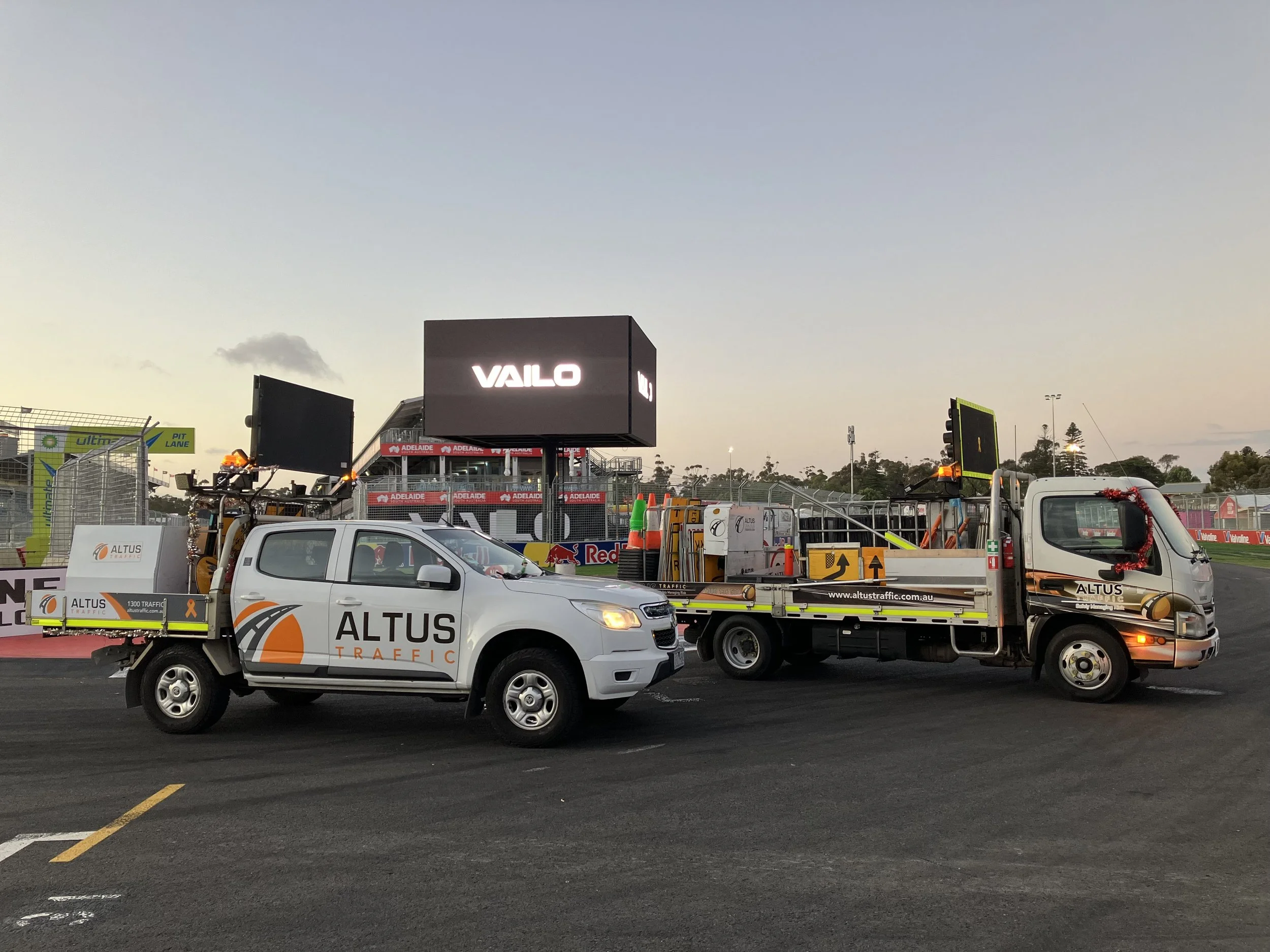 Two traffic service trucks parked on a racetrack near a scoreboard, one marked 'ALTUS TRAFFIC' and the other with a Christmas wreath on the front.