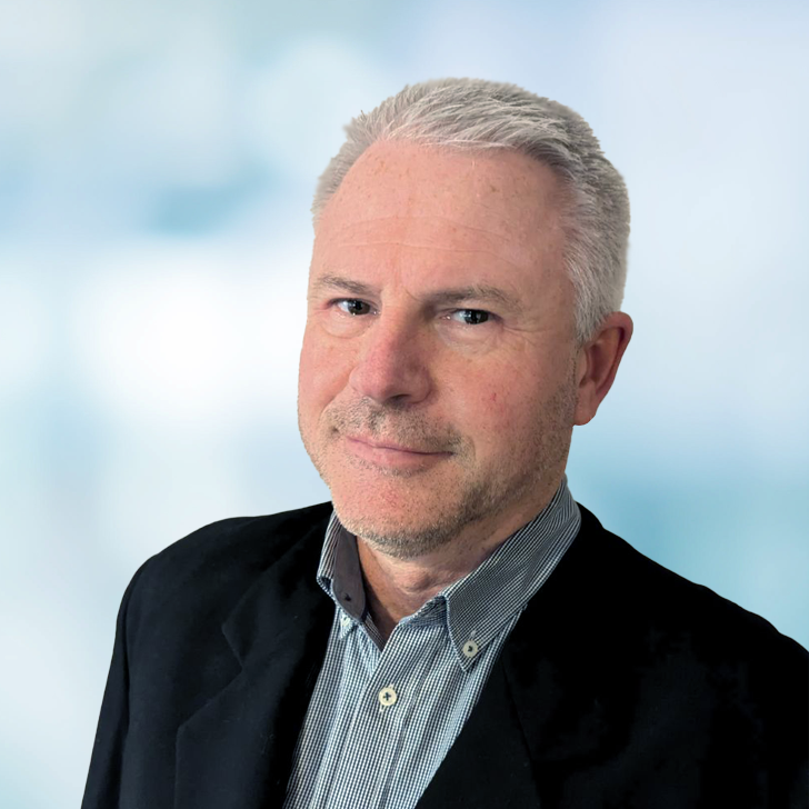 A middle-aged man with short gray hair, wearing a dark blazer over a button-up shirt, looking at the camera against a blurred light blue background.