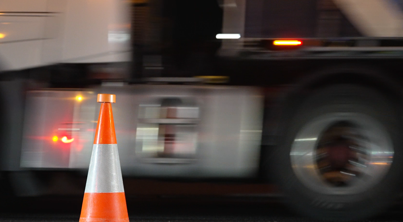 Orange traffic cone with white reflective stripe in front of a moving vehicle or truck.