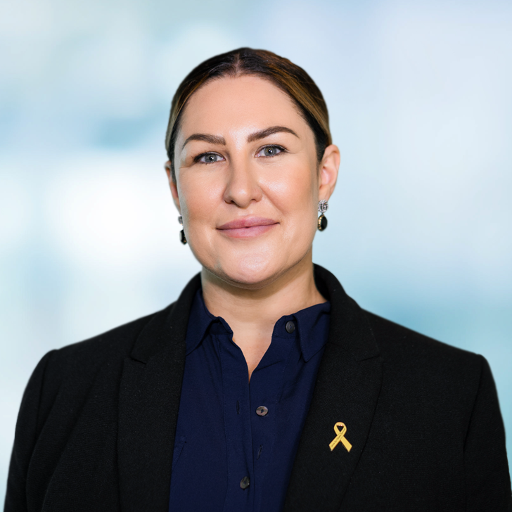 A woman with brown hair, pale skin, blue eyes, and a confident smile wearing a black blazer, navy shirt, and a yellow ribbon pin, standing against a blurred light blue background.