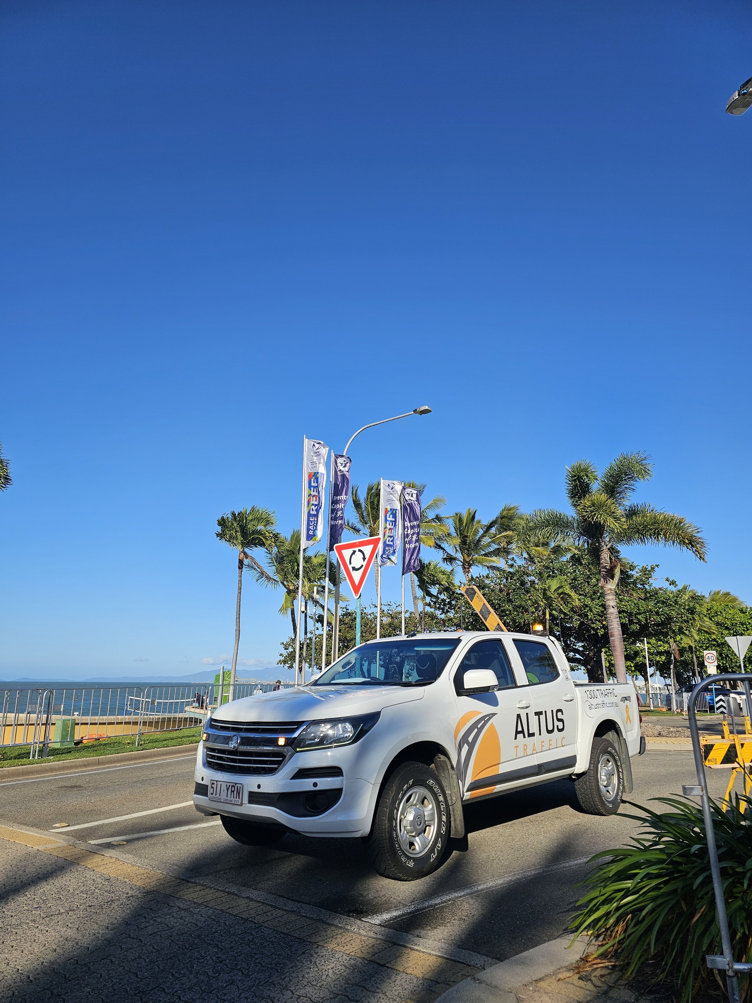 White ALTUS traffic control vehicle parked on a road near palm trees, with traffic signs and banners, under a clear blue sky.