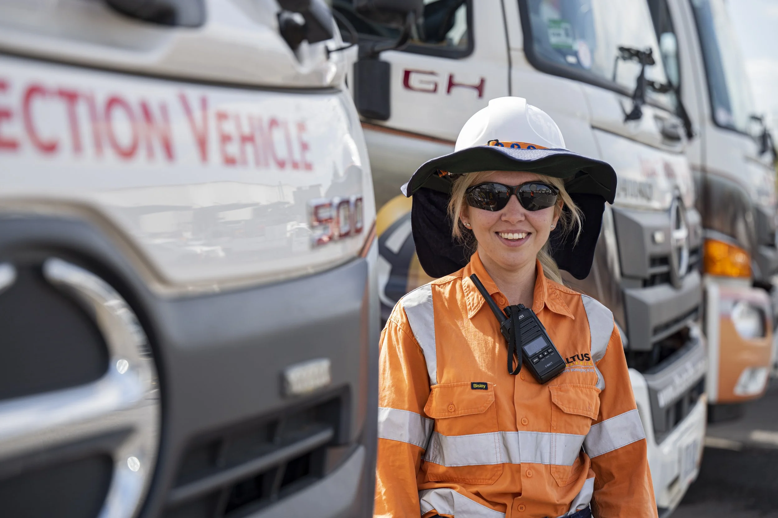 Smiling woman in orange safety vest, wide-brim hat, and sunglasses standing among large trucks labeled 'Protection Vehicle' at a work site.