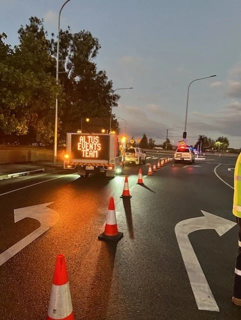 Road construction or accident scene with traffic cones, emergency vehicle lights, and a sign reading 'ALTUS EVENTS TEAM' on a digital display.