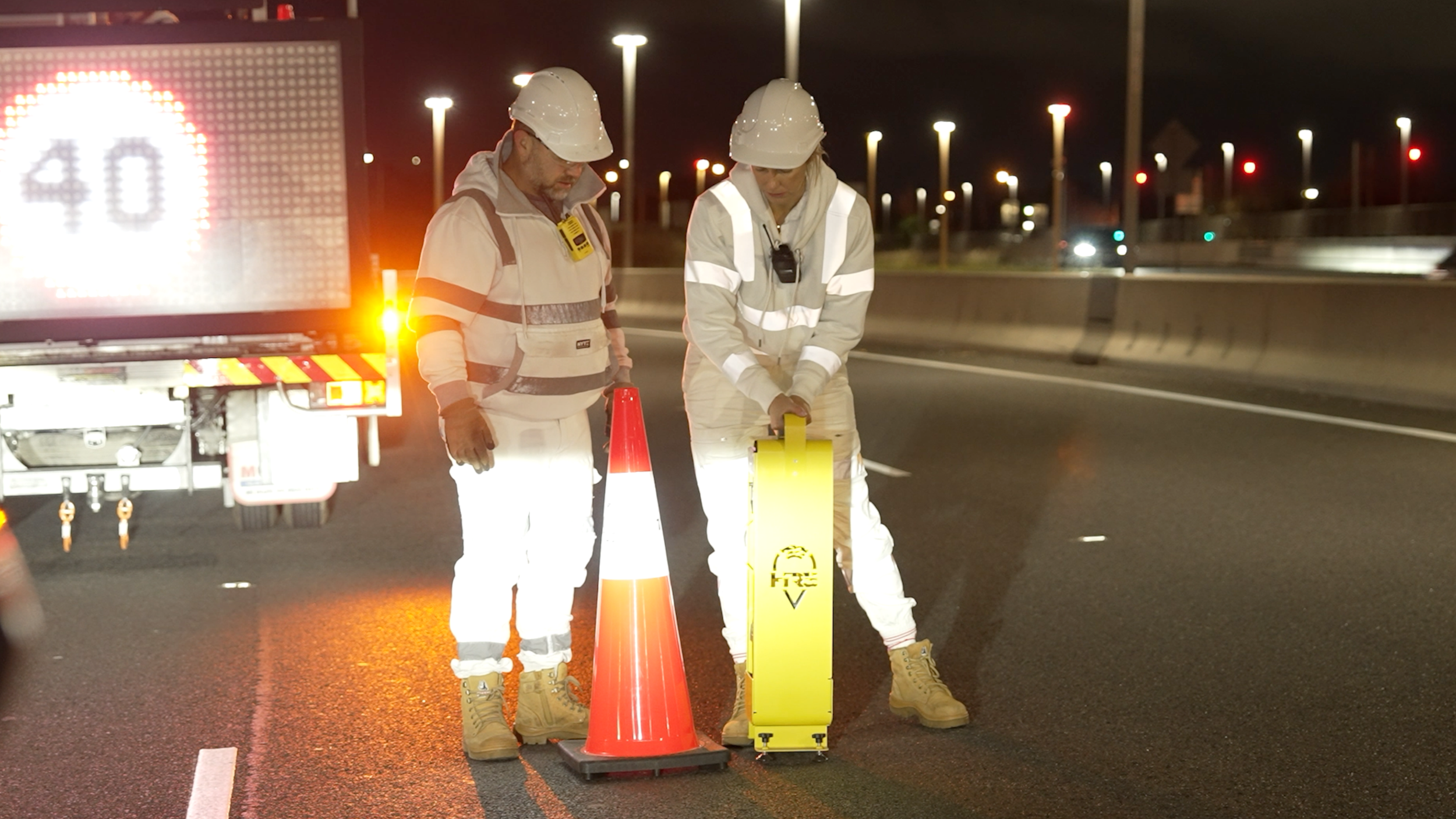 Two workers in safety gear inspect the road at night near a maintenance vehicle with flashing lights, cones, and a yellow equipment case.
